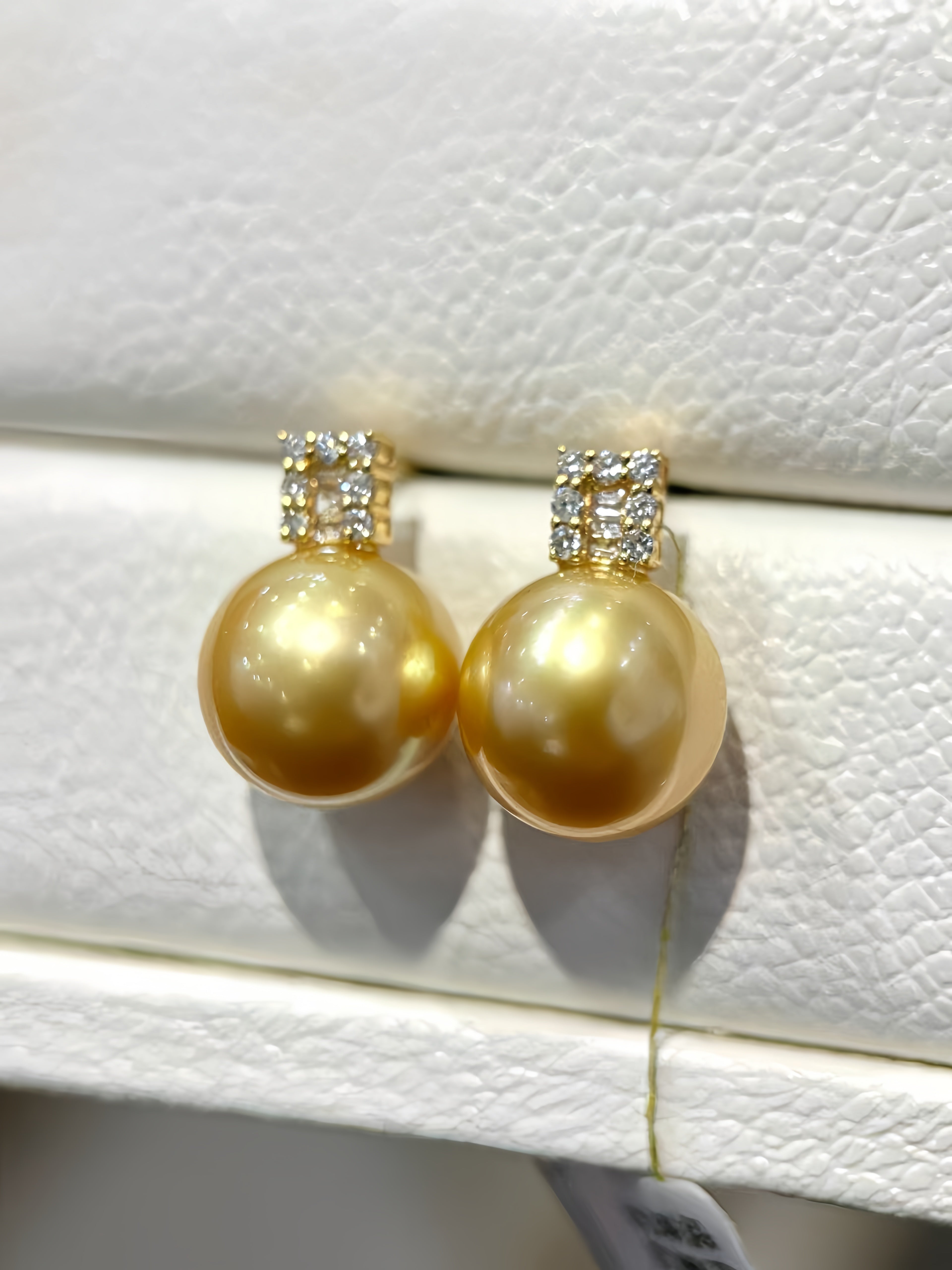 A pair of gold stud earrings featuring large round gold freshwater pearls and a small square cluster of clear stones at the top, displayed on a textured white jewelry stand.