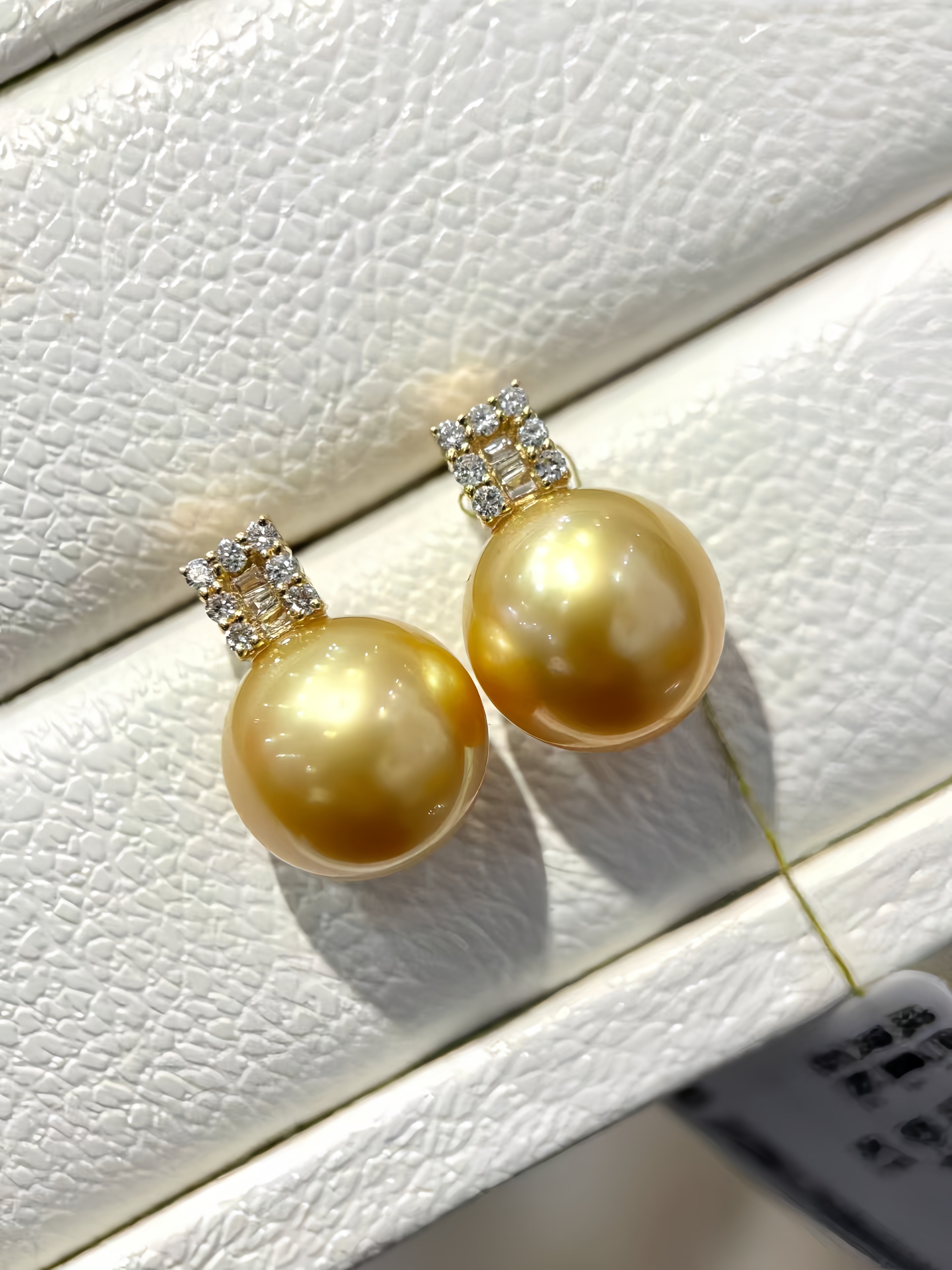 A pair of gold freshwater pearl drop earrings with small diamond-set crowns, displayed on a textured white jewelry box.