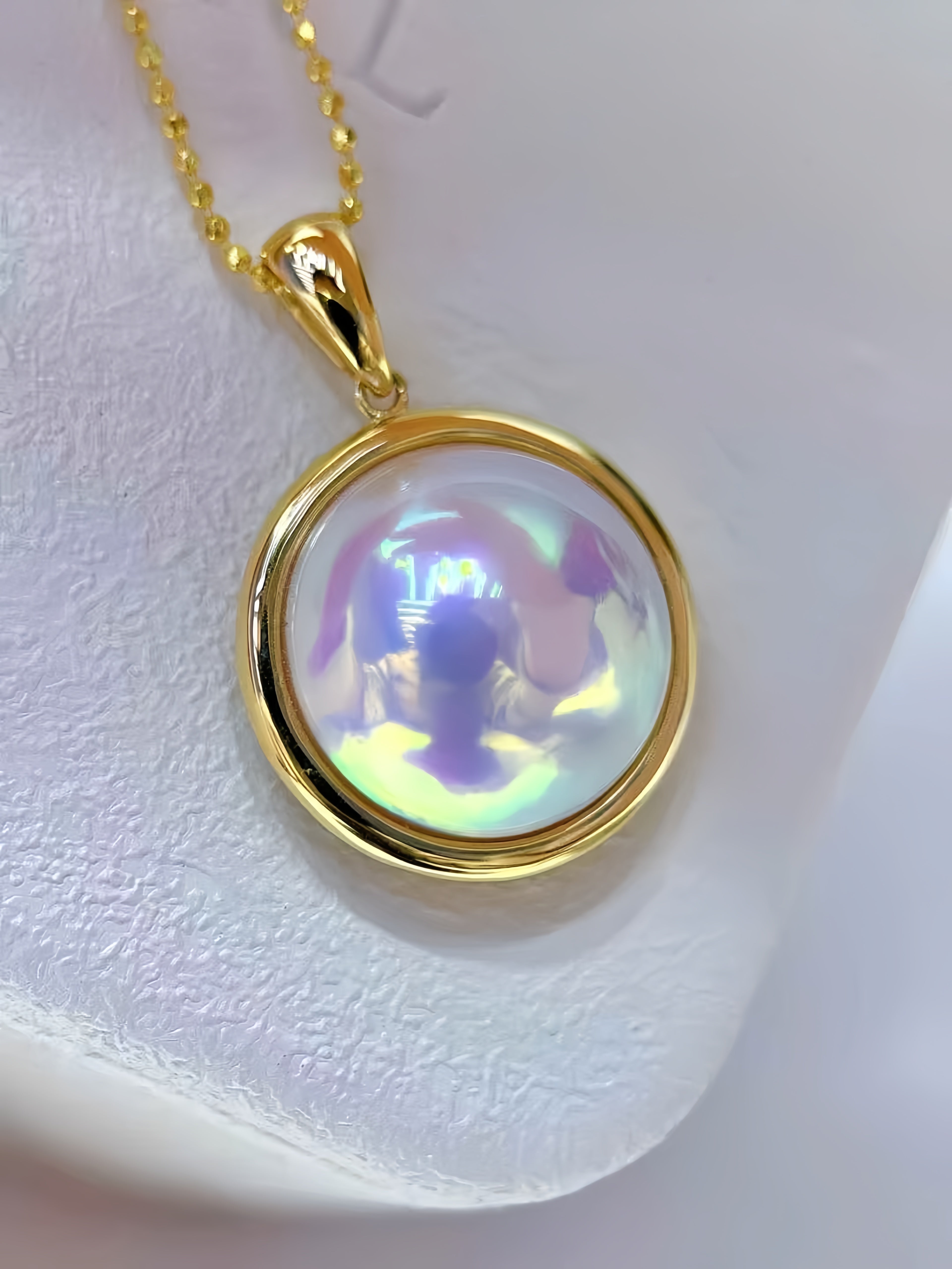 A gold pendant necklace with a round iridescent shell inlay, displayed on a textured white surface.