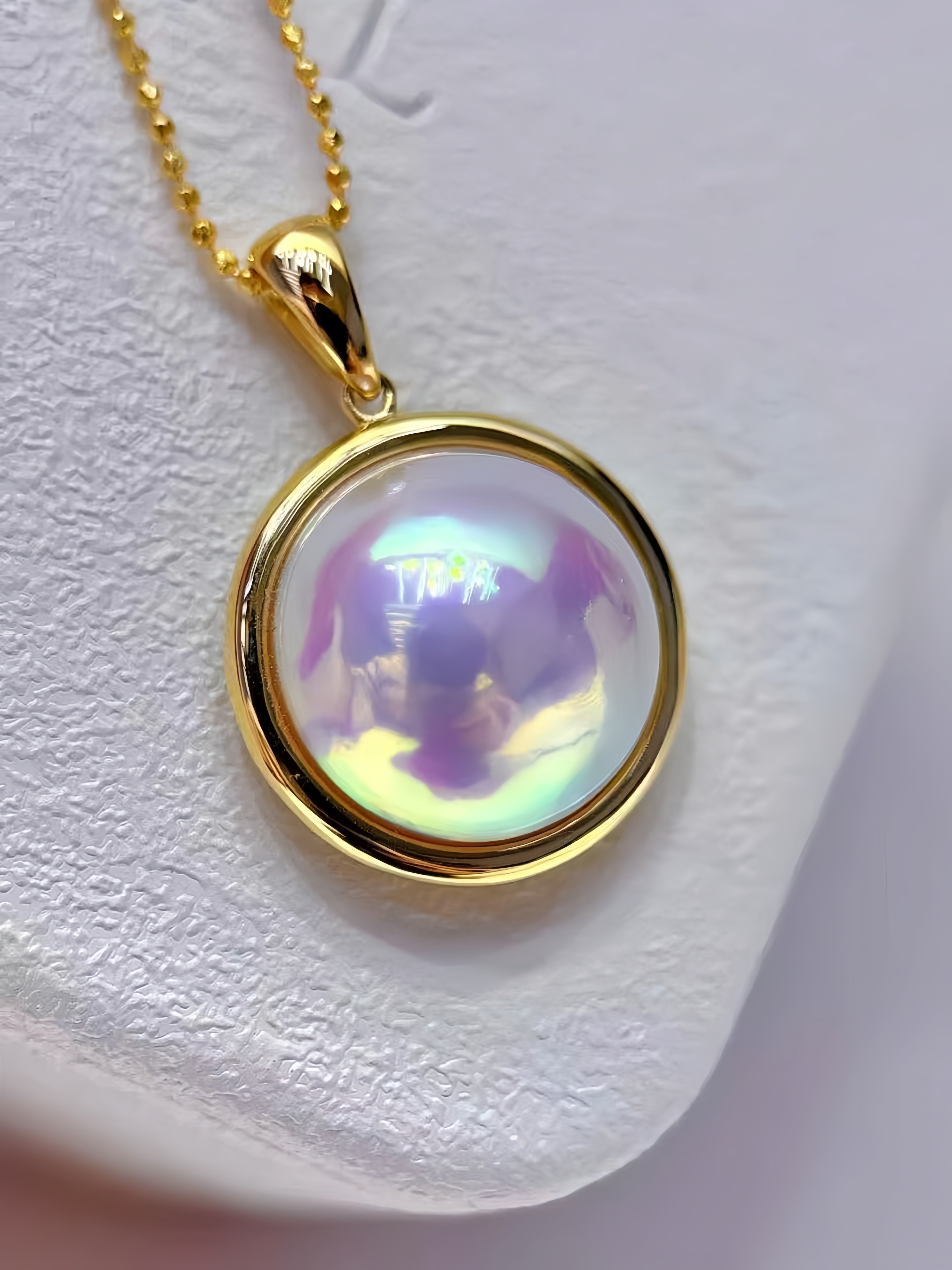 A gold pendant necklace with a round bezel containing an iridescent, milky white stone that reflects purple and green hues, resting on a textured white surface.