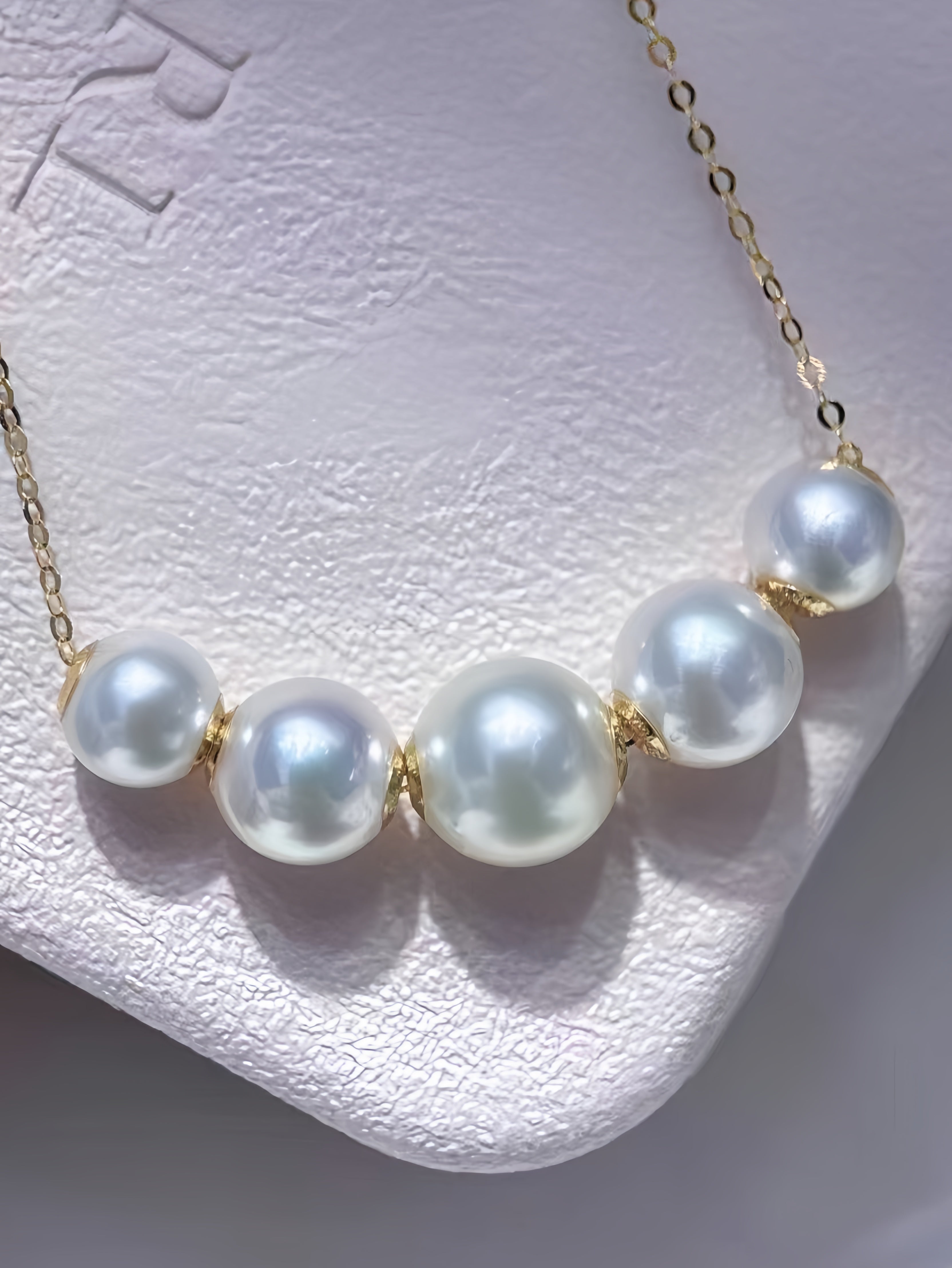 A close-up of a necklace with five large white pearls and gold chain links, displayed on a textured white jewelry stand.