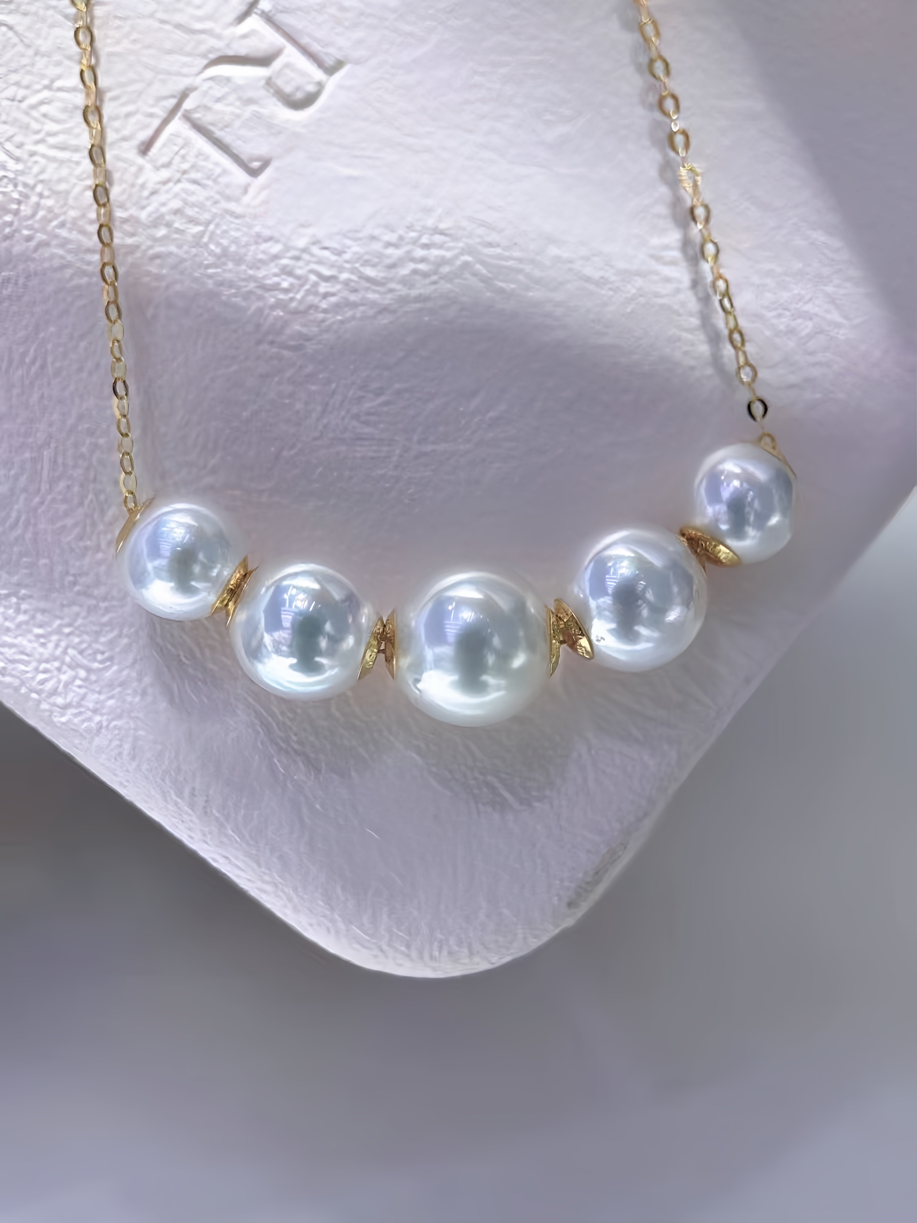 A gold chain necklace featuring five large, lustrous white freshwater pearls spaced along the chain, displayed on a textured white jewelry stand.