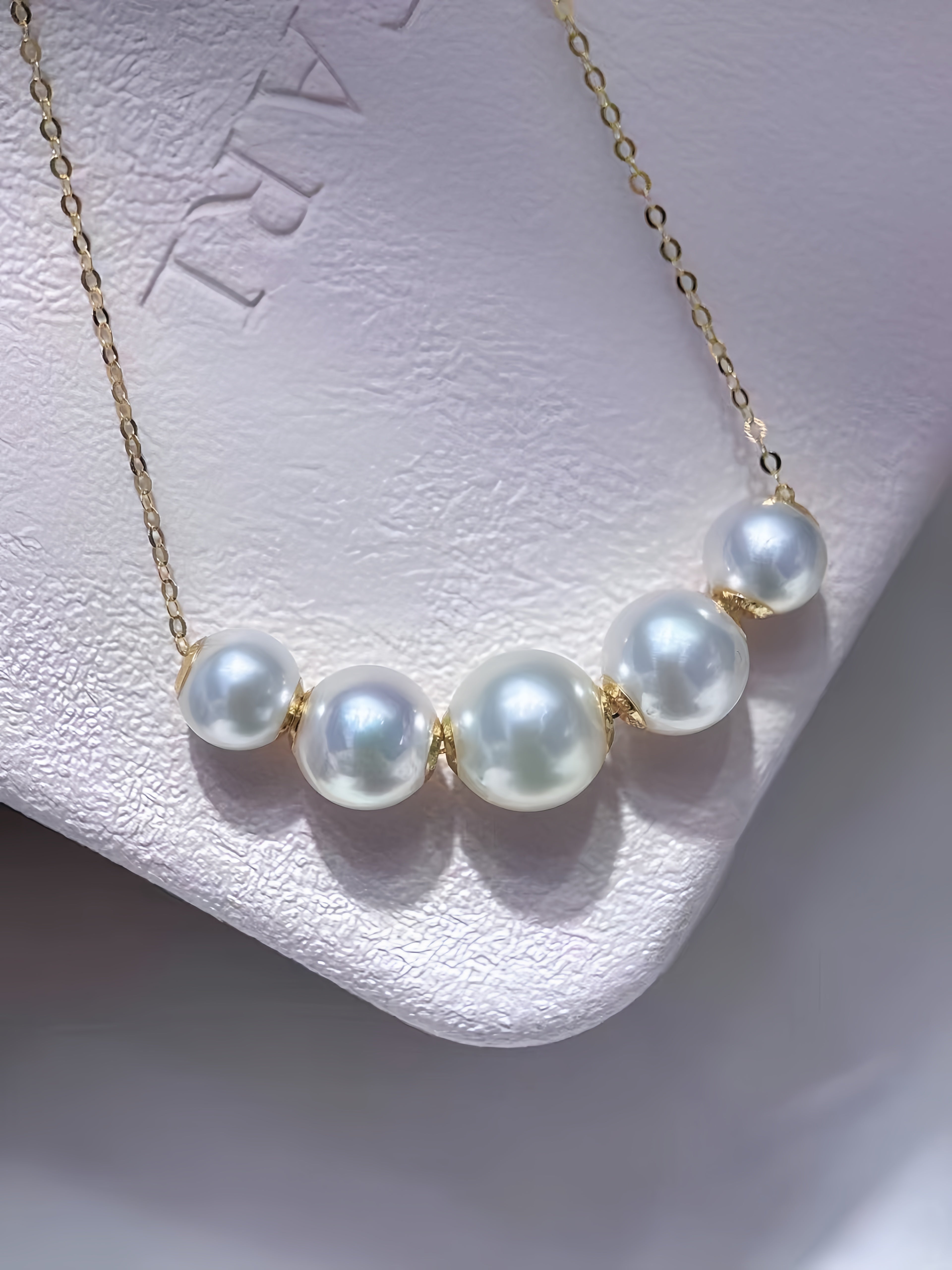 A gold chain necklace featuring five lustrous white freshwater pearls spaced along the front, displayed on a textured white jewelry stand.