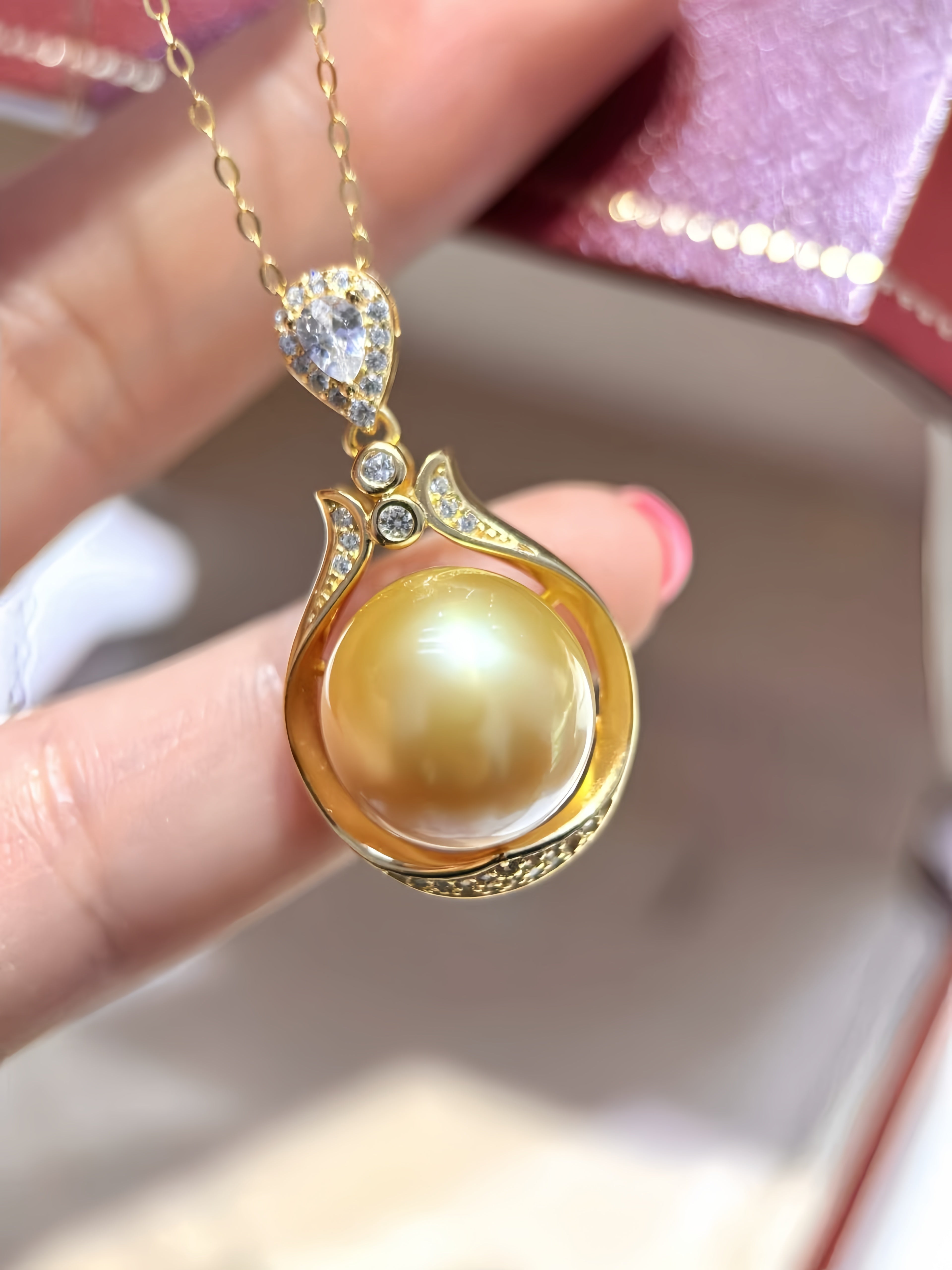 A gold pendant necklace featuring a large round yellow pearl in a decorative gold setting, held by a hand. The pendant has small clear accent stones and is shown against a soft-focus background with a pinkish box.