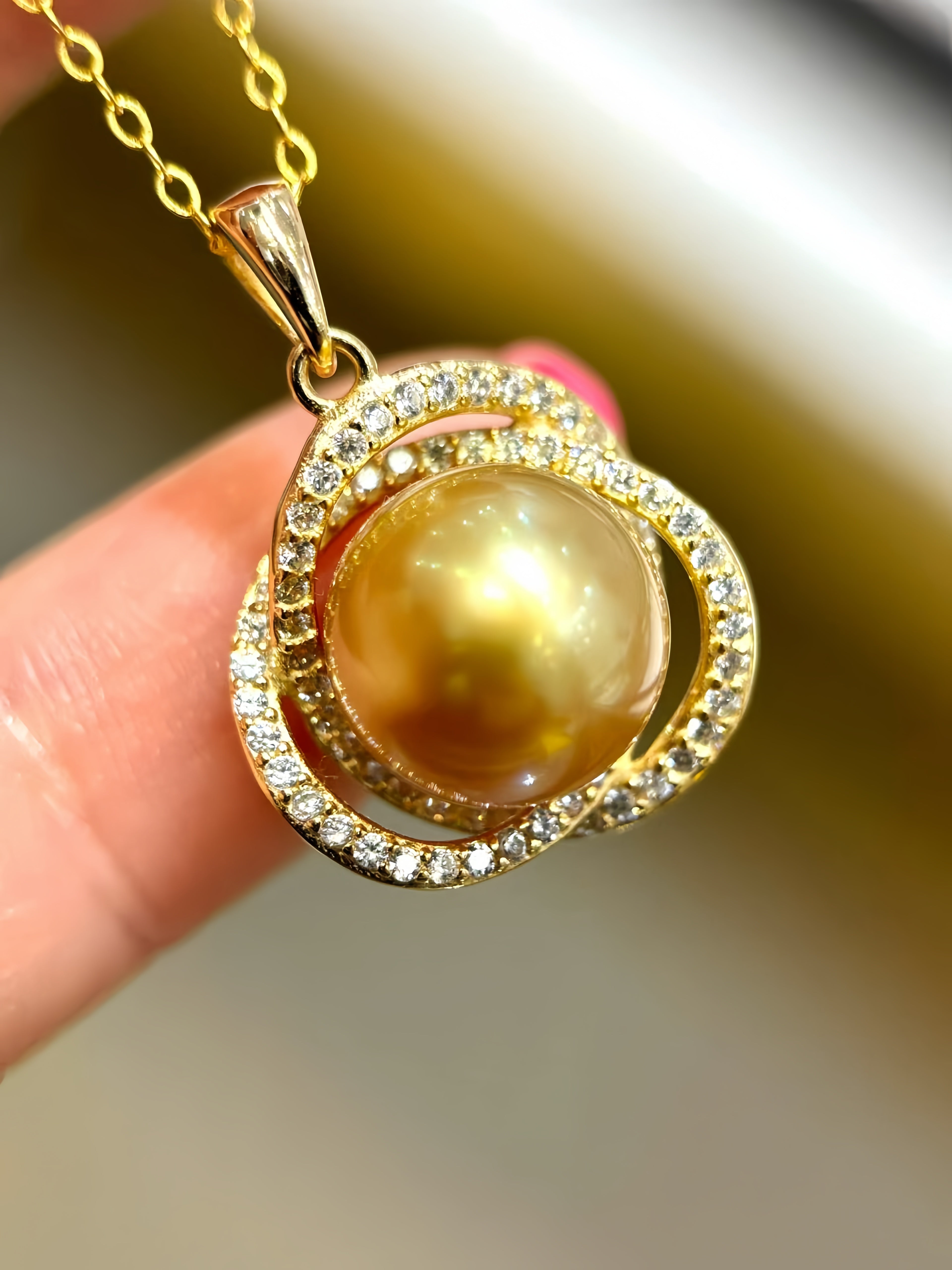 A gold pendant necklace featuring a large round golden pearl surrounded by double rings of small clear crystals, held by a hand against a blurred background.