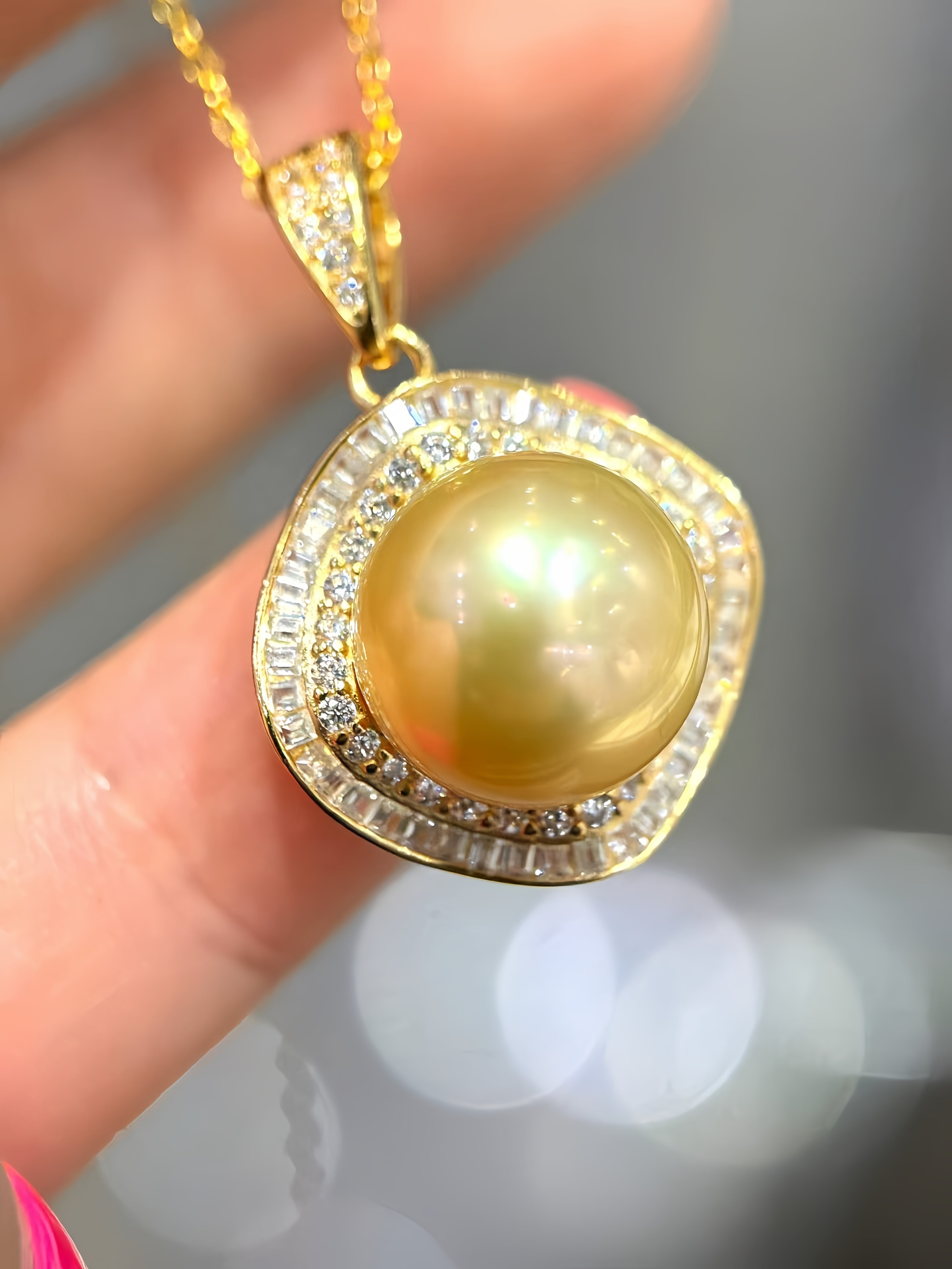 A gold pendant necklace featuring a large round yellow pearl surrounded by a halo of small clear stones, held between fingers against a soft blurred gray background.