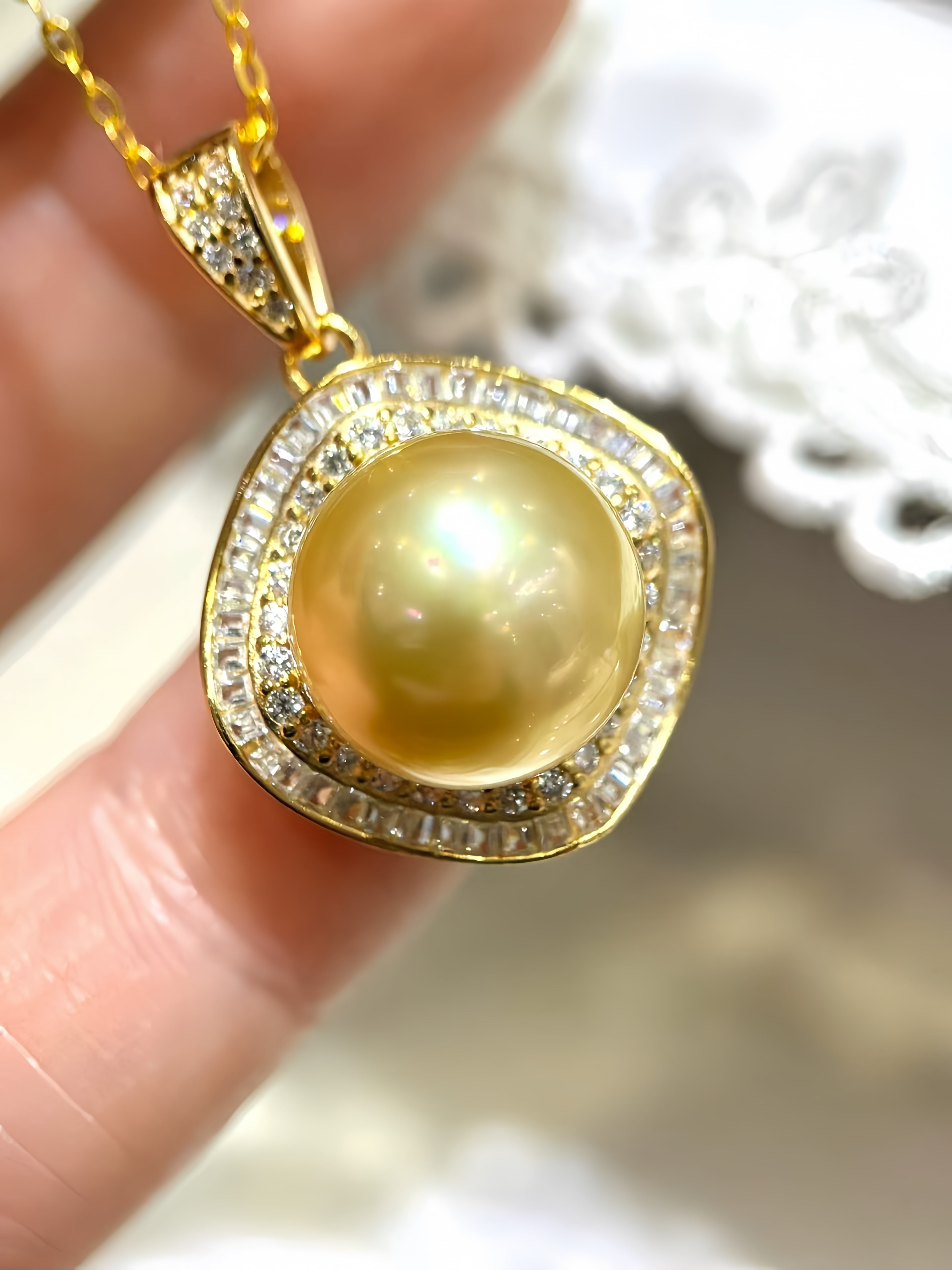A gold pendant necklace featuring a large round yellow pearl surrounded by a double halo of small clear stones, held between fingers against a soft white lace background.