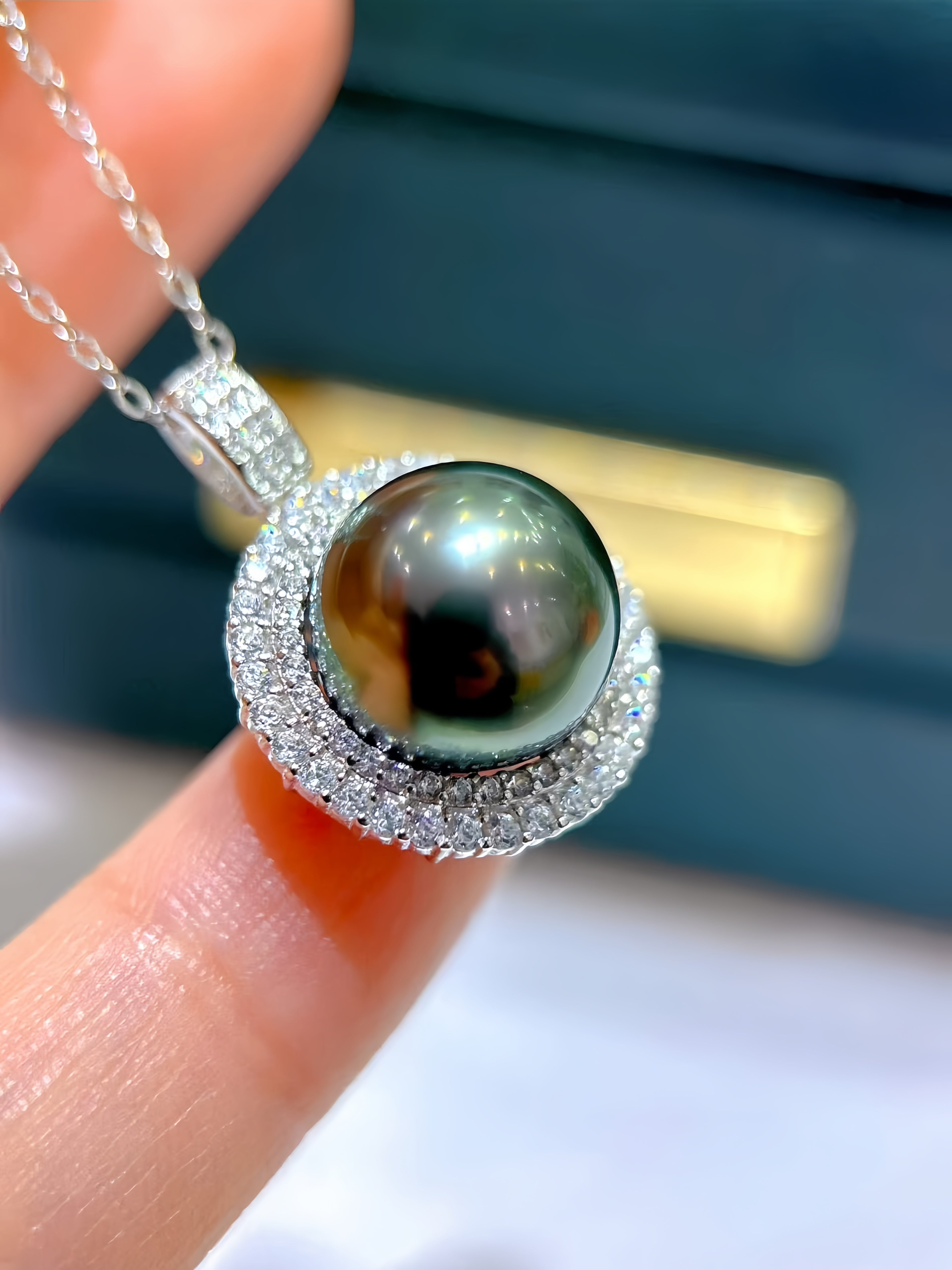 A close-up of a silver pendant necklace featuring a large, round, dark green-black Tahitian pearl surrounded by a halo of small clear stones, held between fingers.