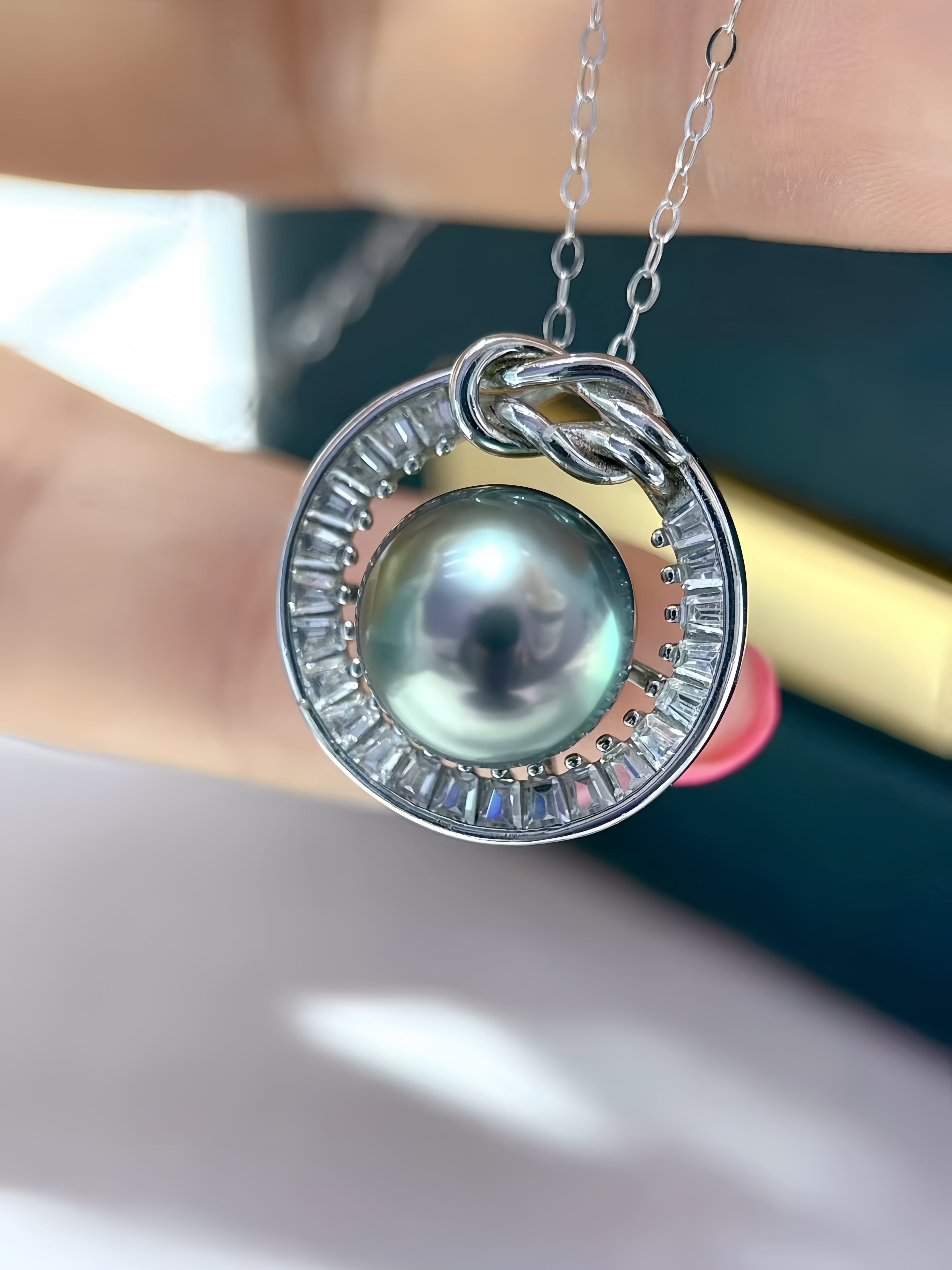 A close-up of a silver pendant necklace featuring a round dark green pearl surrounded by clear baguette crystals, held by a hand.
