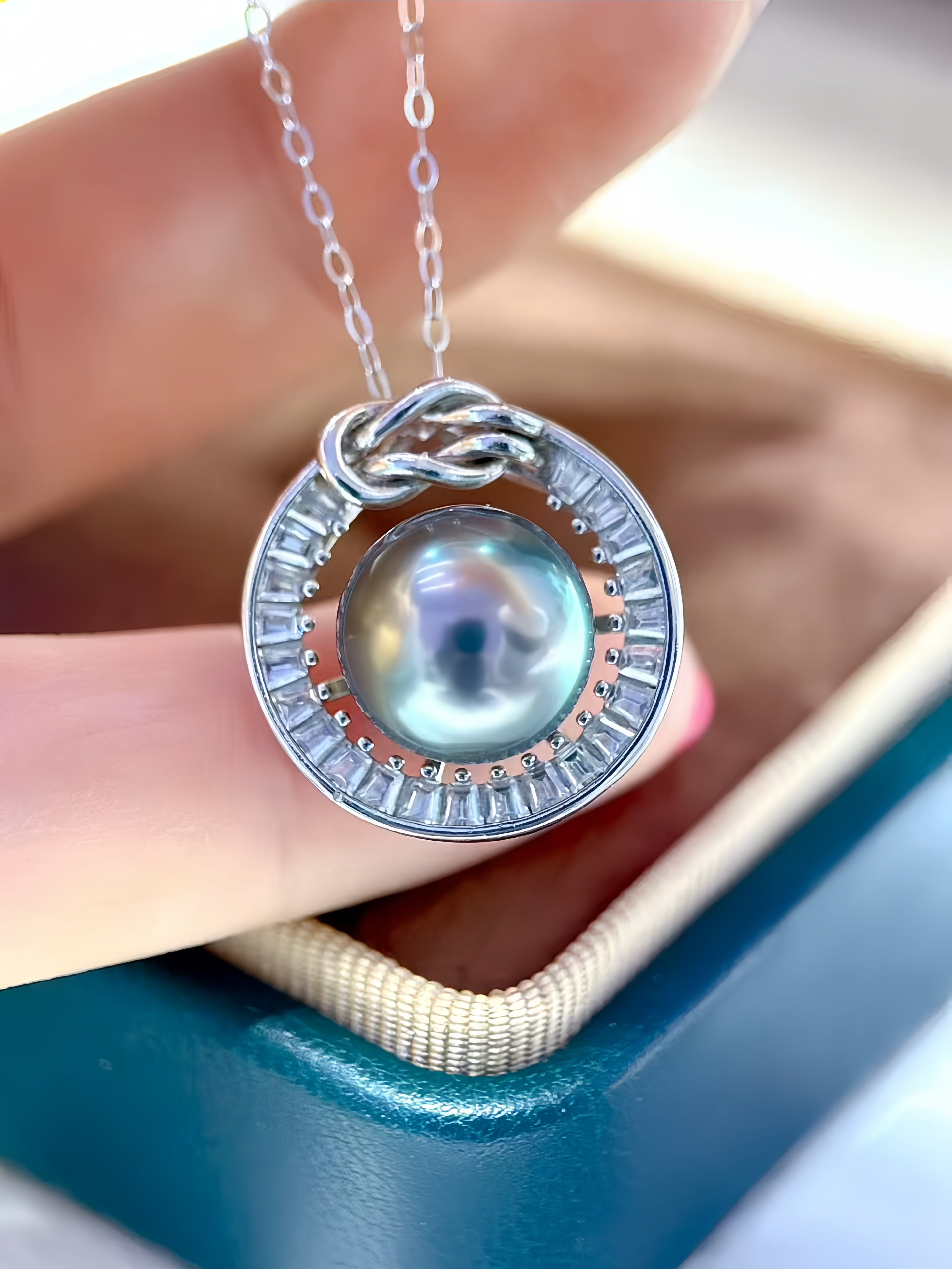 A sterling silver pendant necklace featuring a central iridescent pearl surrounded by a halo of small clear stones and a knot detail at the top, held by a hand over a teal jewelry box.