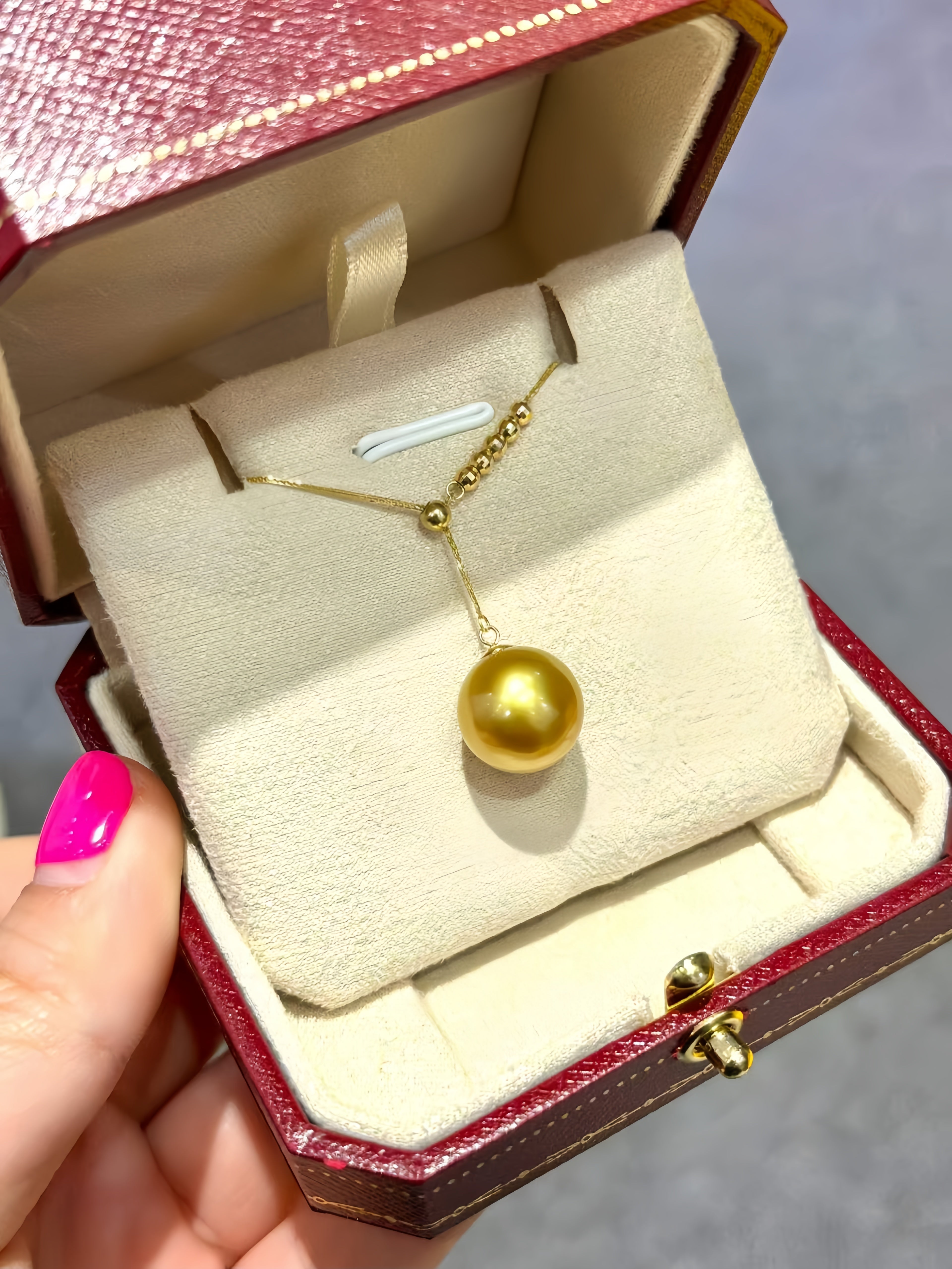 A gold pendant necklace with a large round pearl-like drop is displayed inside an open red jewelry box with a cream velvet insert. A hand with bright pink nail polish holds the box on the left.