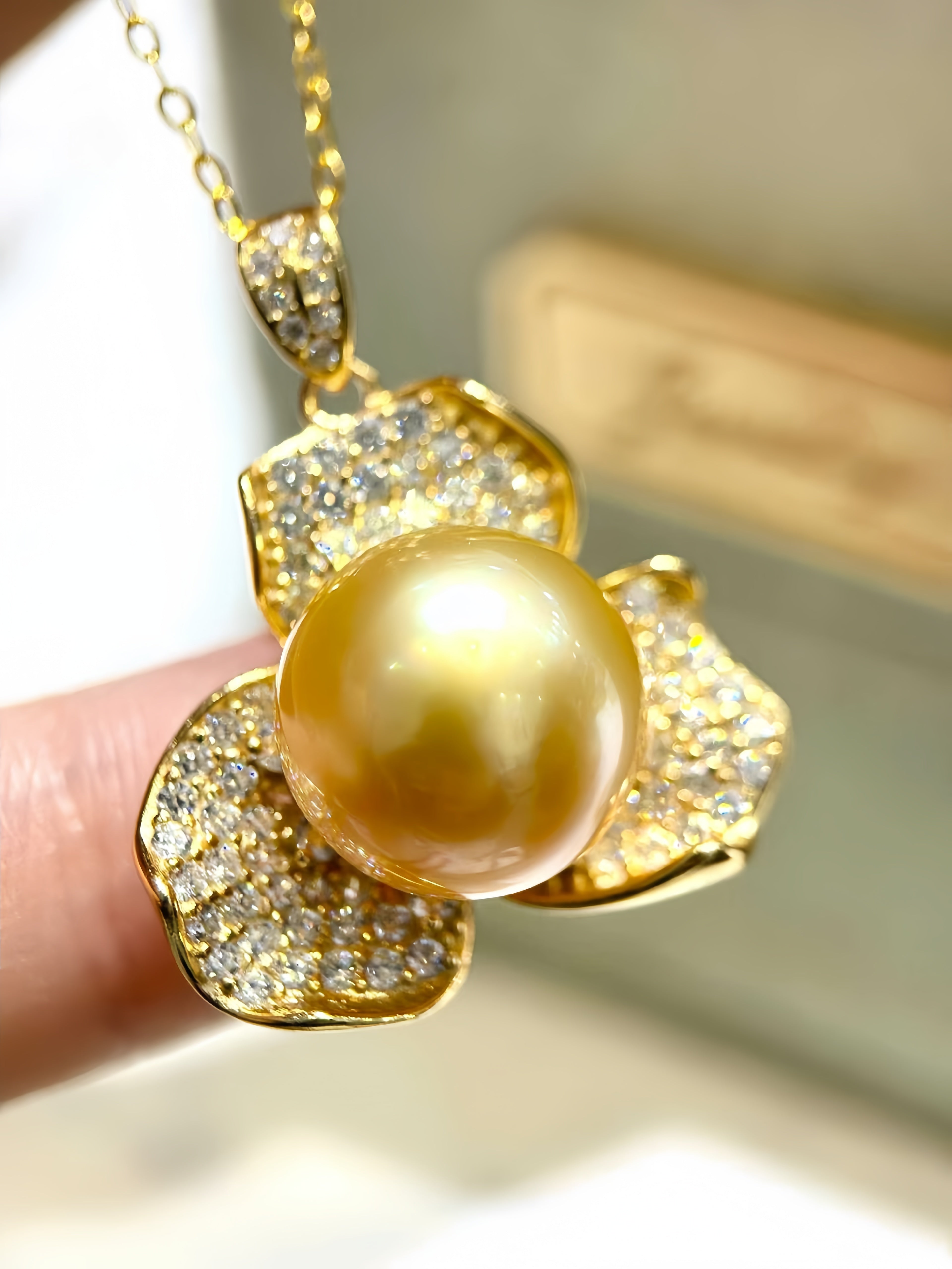 A gold pendant necklace featuring a large round golden pearl surrounded by flower-shaped settings paved with small clear stones, held by a finger against a softly blurred background.