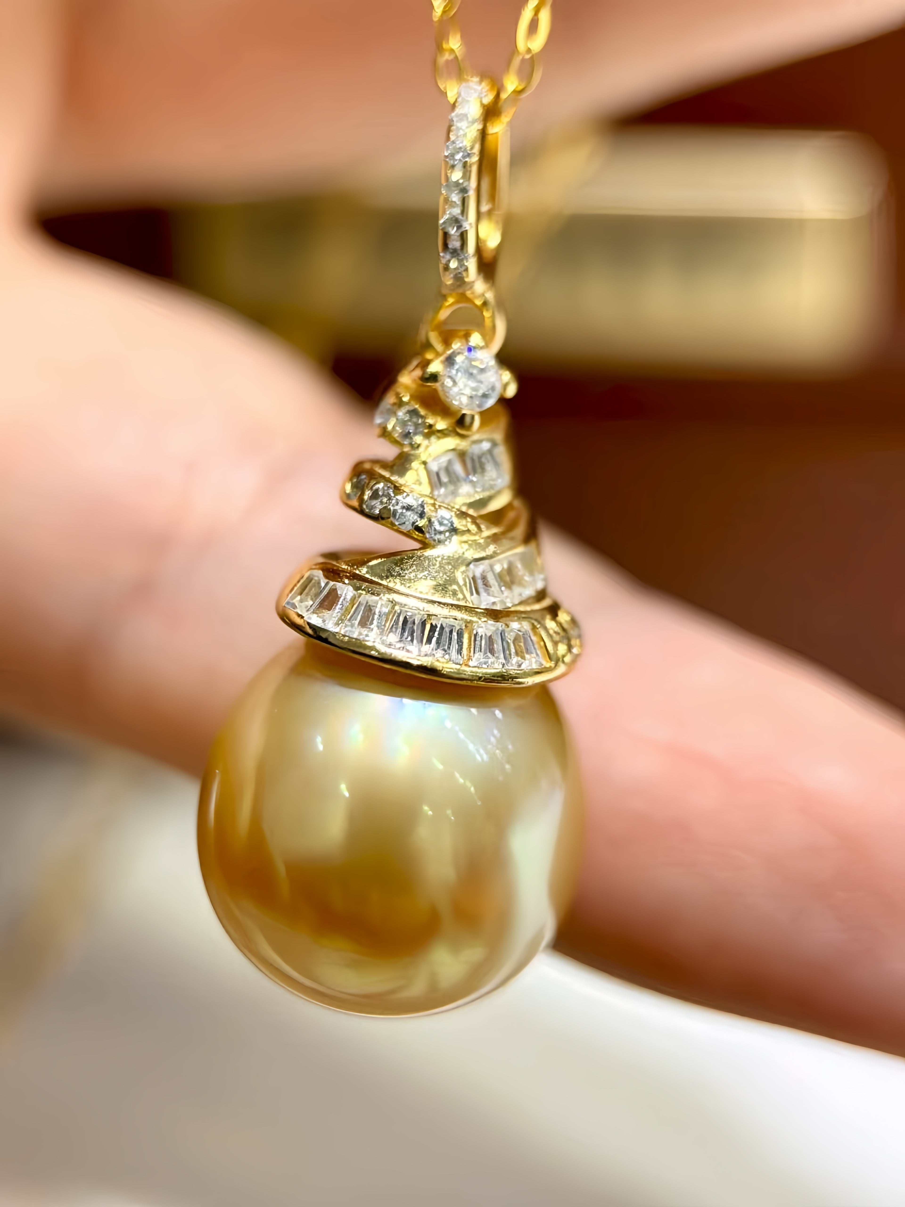A gold pendant necklace featuring a large, lustrous pearl-like bead with a spiral arrangement of small clear stones, held against a blurred background.