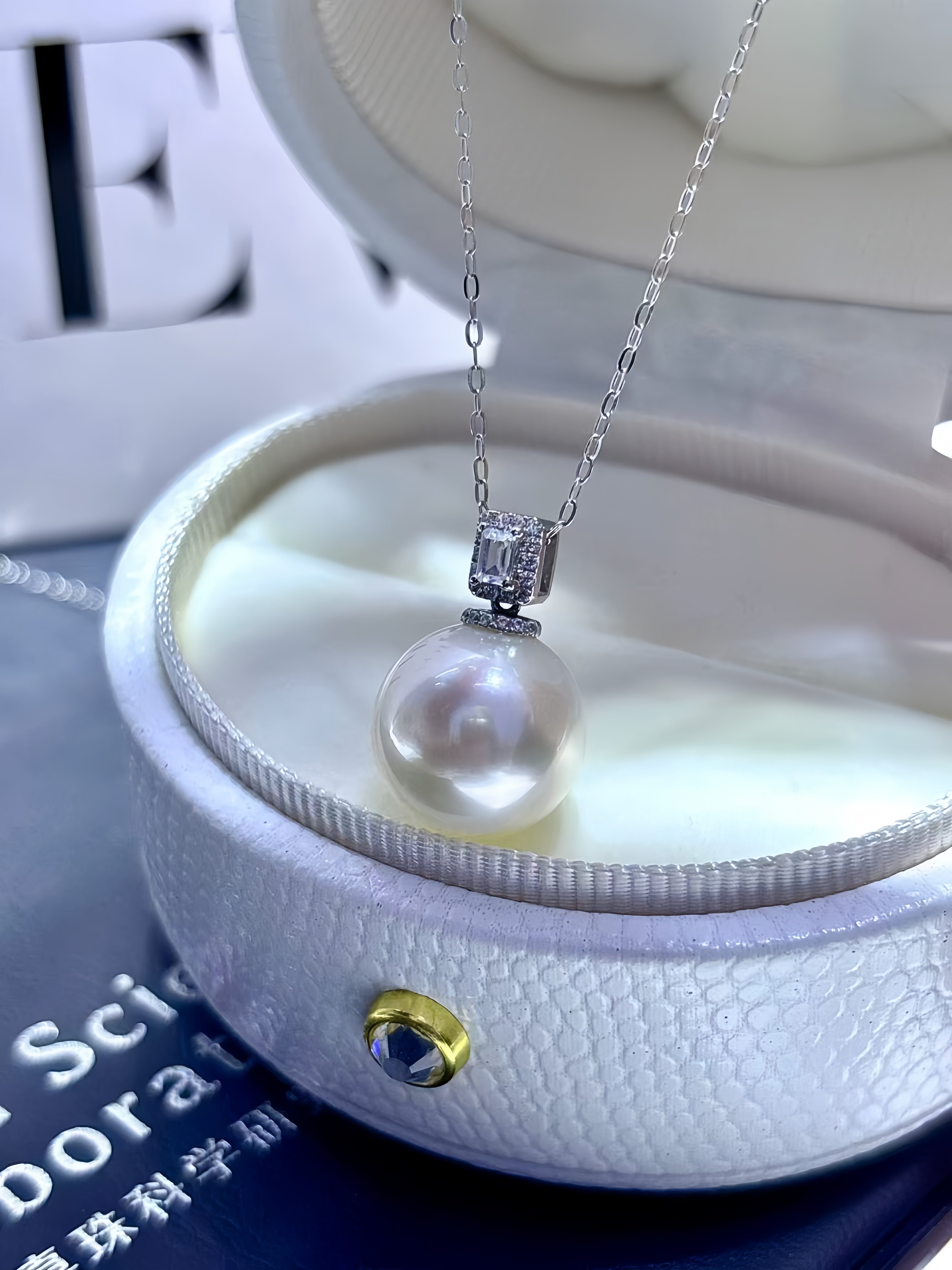 A silver chain necklace with a rectangular bezel-set clear stone pendant and a large round white pearl drop, displayed inside an open white jewelry box.