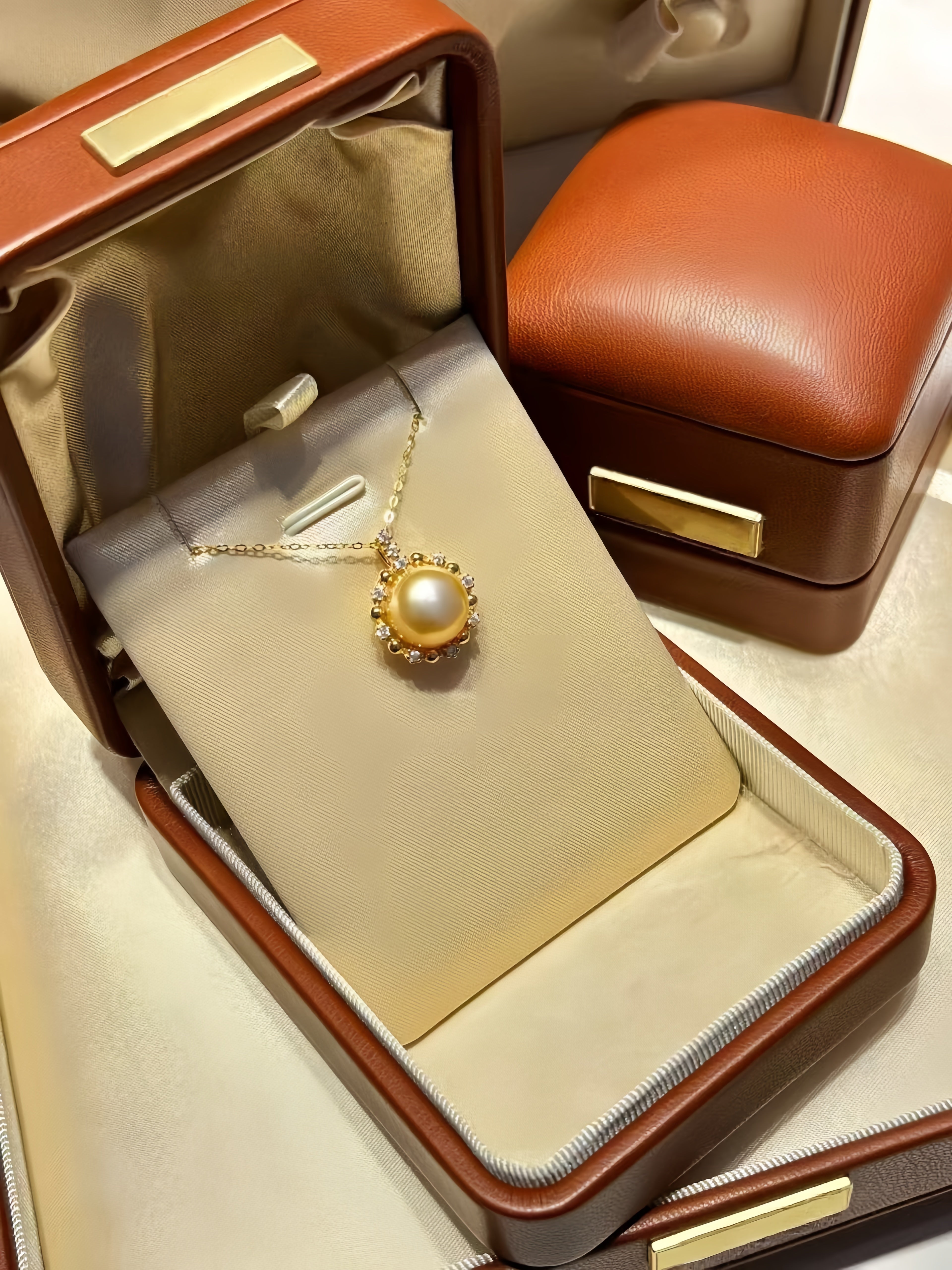 A gold pendant necklace with a large white pearl is displayed inside an open brown leather jewelry box with gold-tone hardware. A second closed matching jewelry box sits behind it on a light cream surface.