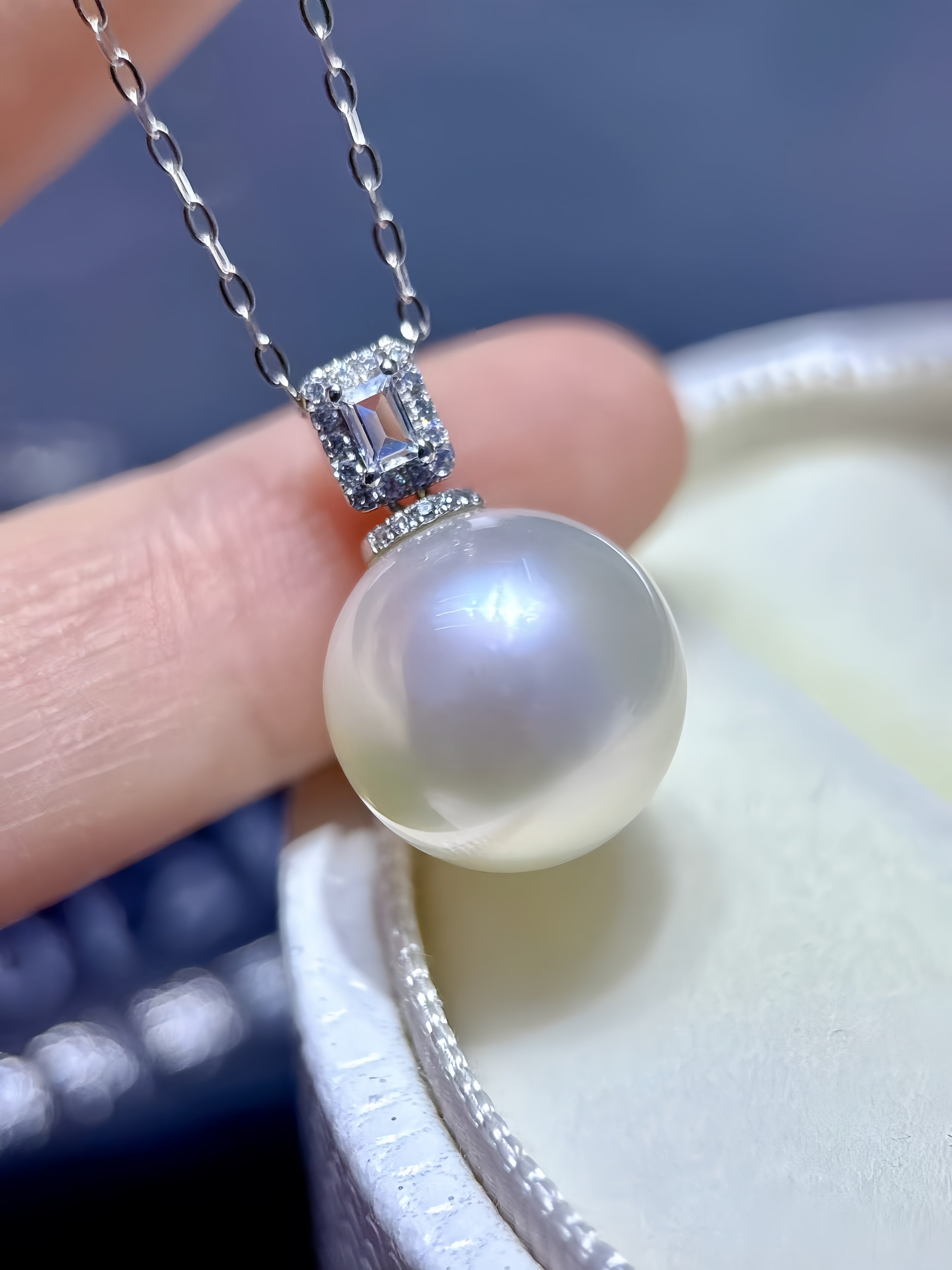 A close-up of a pearl pendant necklace held by a hand, featuring a large round white pearl suspended from a silver-toned bezel set with small clear stones, with a jewelry box in the background.