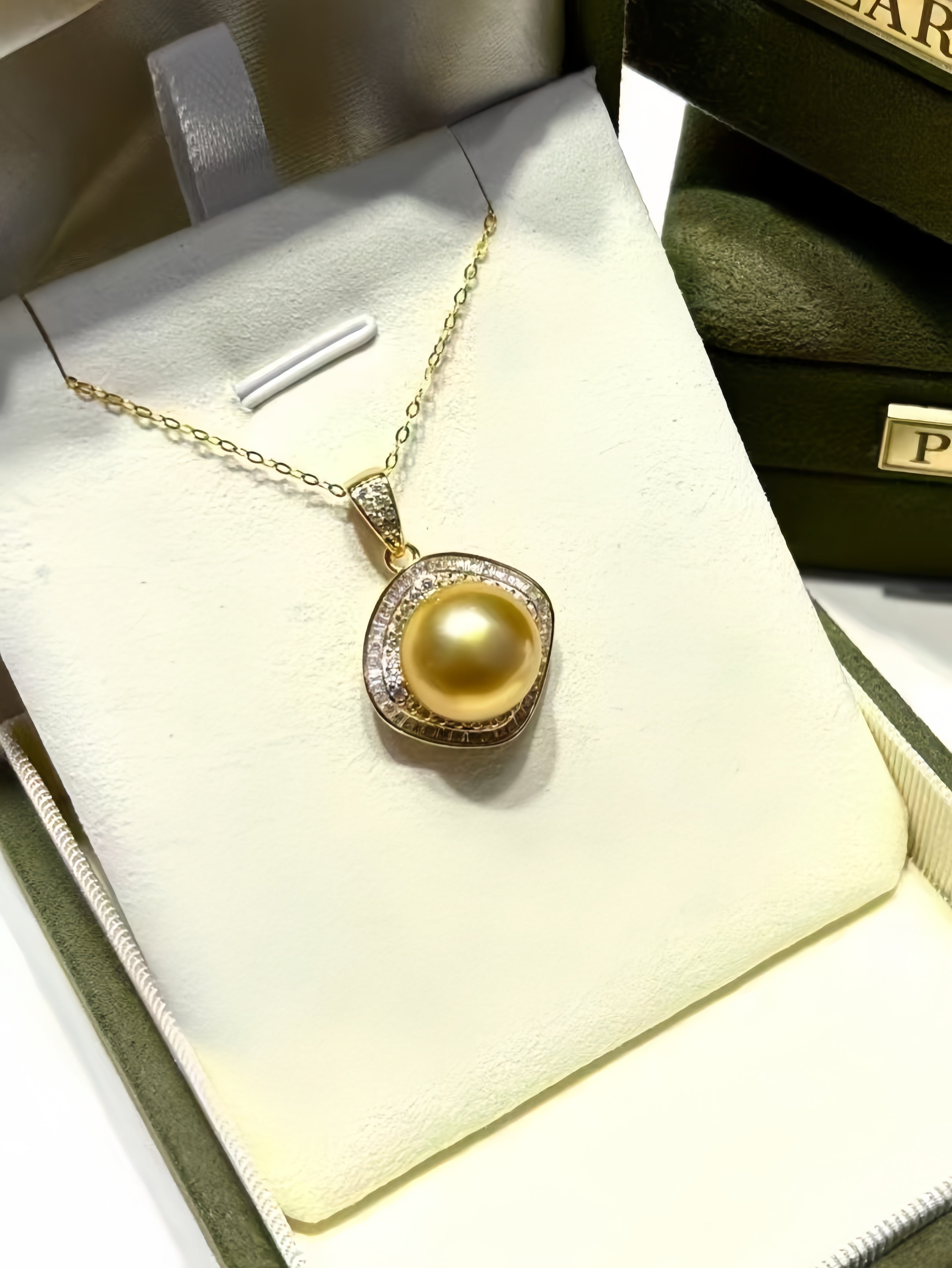 A gold pendant necklace with a large round yellow pearl surrounded by small clear stones, displayed on a cream jewelry cushion inside a dark green box.