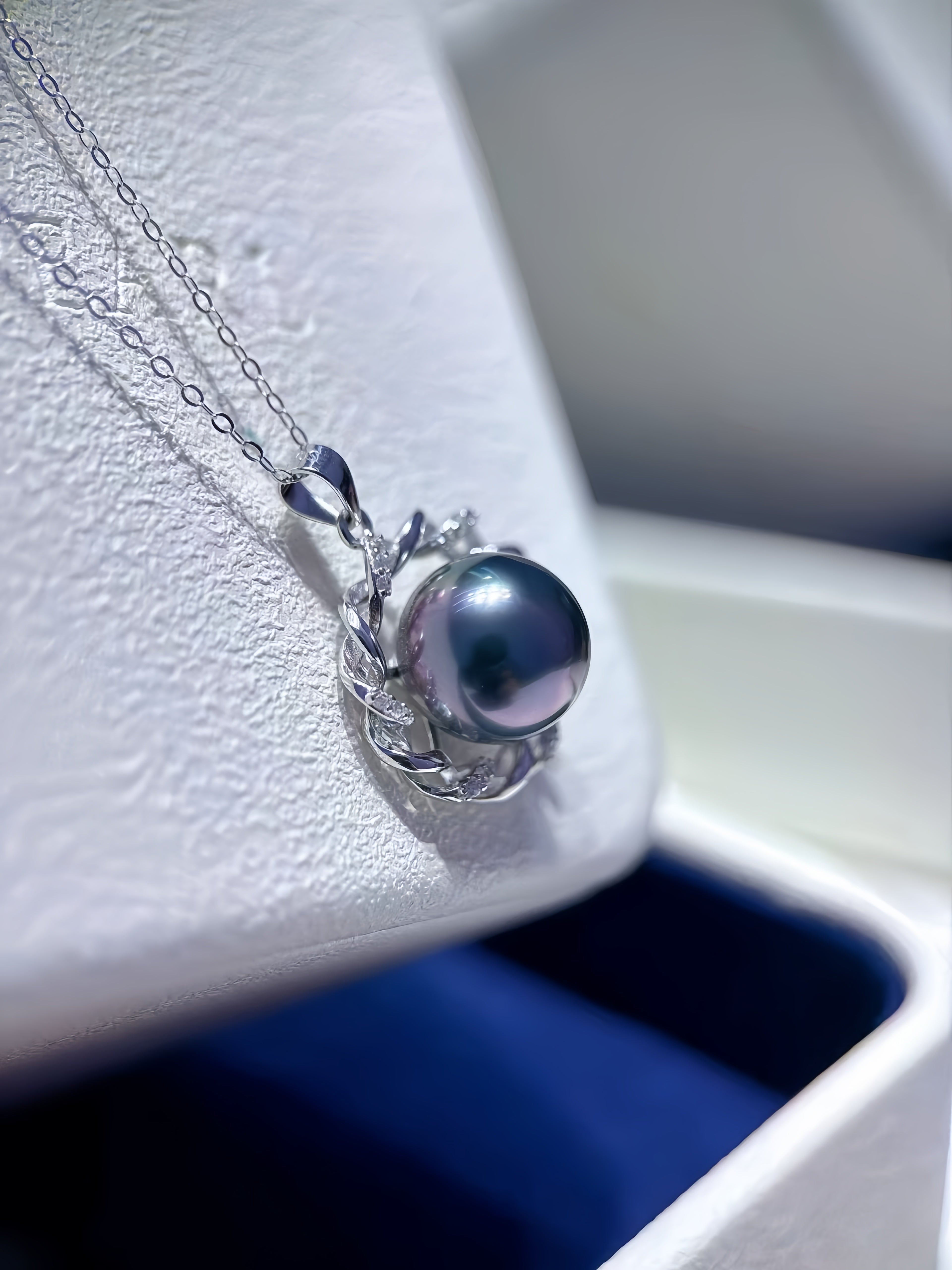 A silver pendant necklace featuring a single dark, lustrous pearl set in a delicate silver filigree frame, resting on a textured white jewelry display stand.