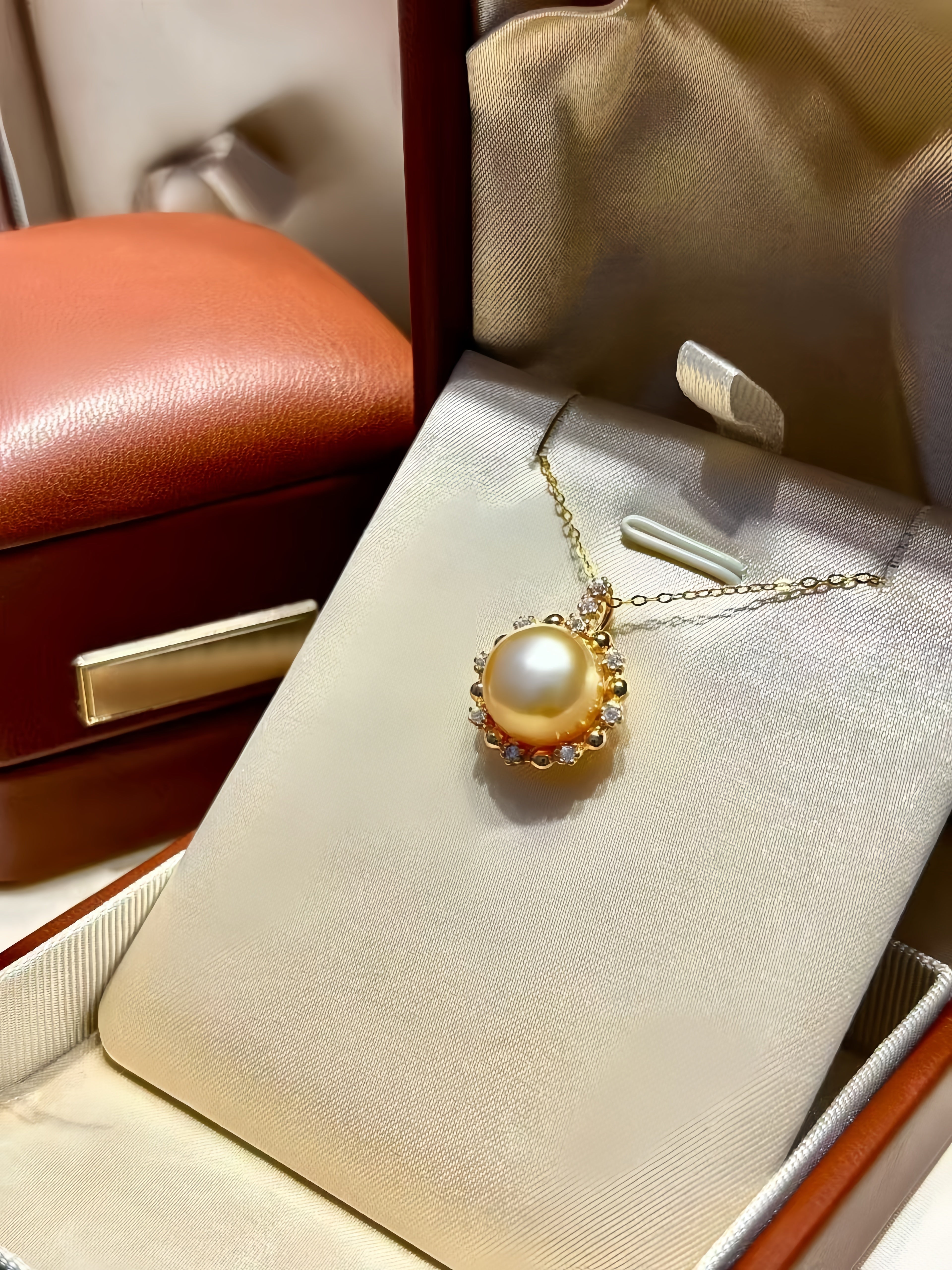 A gold pendant necklace with a large round pearl and small clear stones, displayed on a cream satin jewelry stand inside an open reddish-brown jewelry box.
