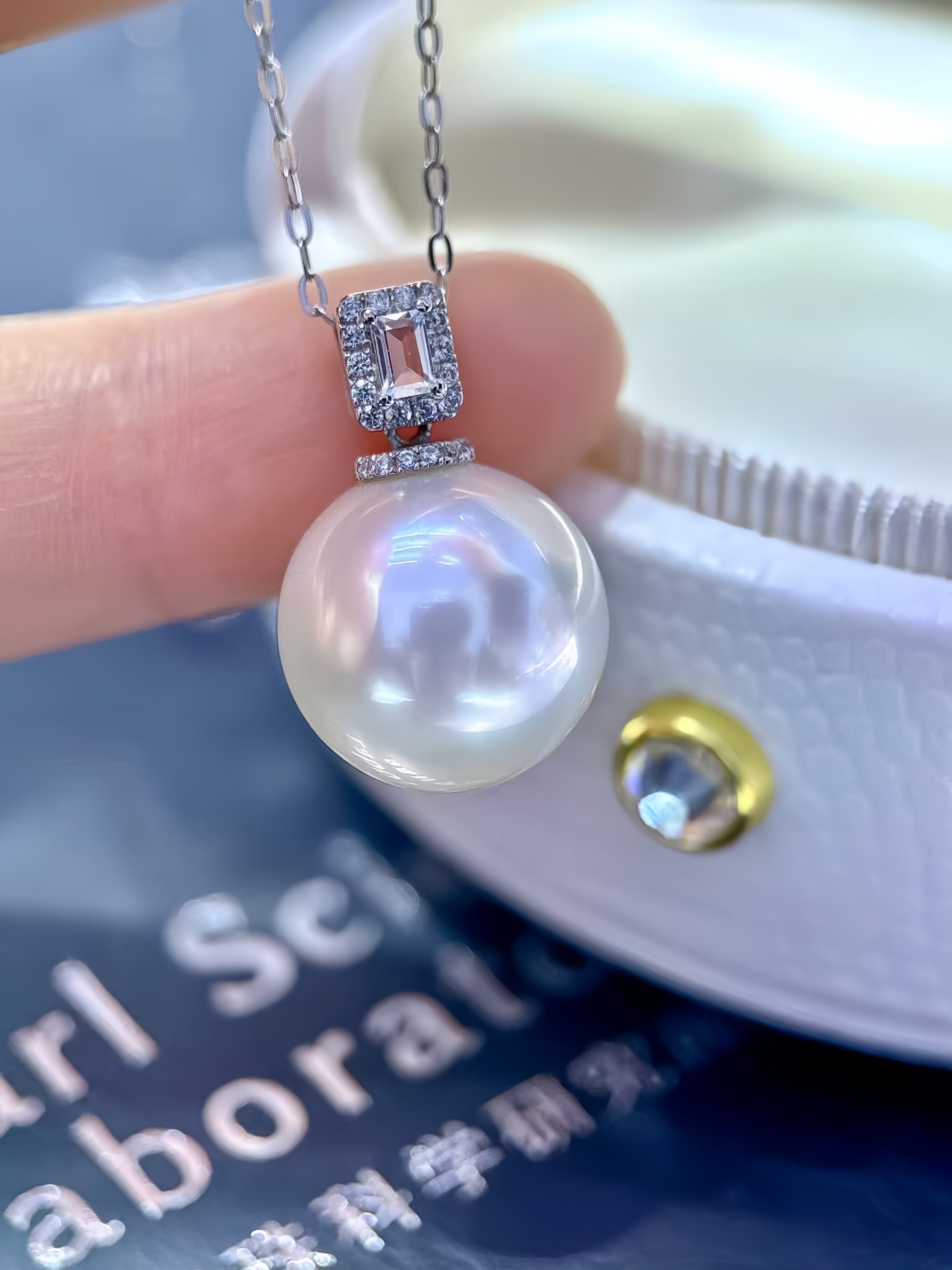 A close-up of a pearl pendant necklace held between fingers, featuring a large round white pearl with iridescent luster, a silver-toned setting with a rectangular clear gemstone, and a delicate chain. A white textured jewelry box and a dark blue printed card are blurred in the background.