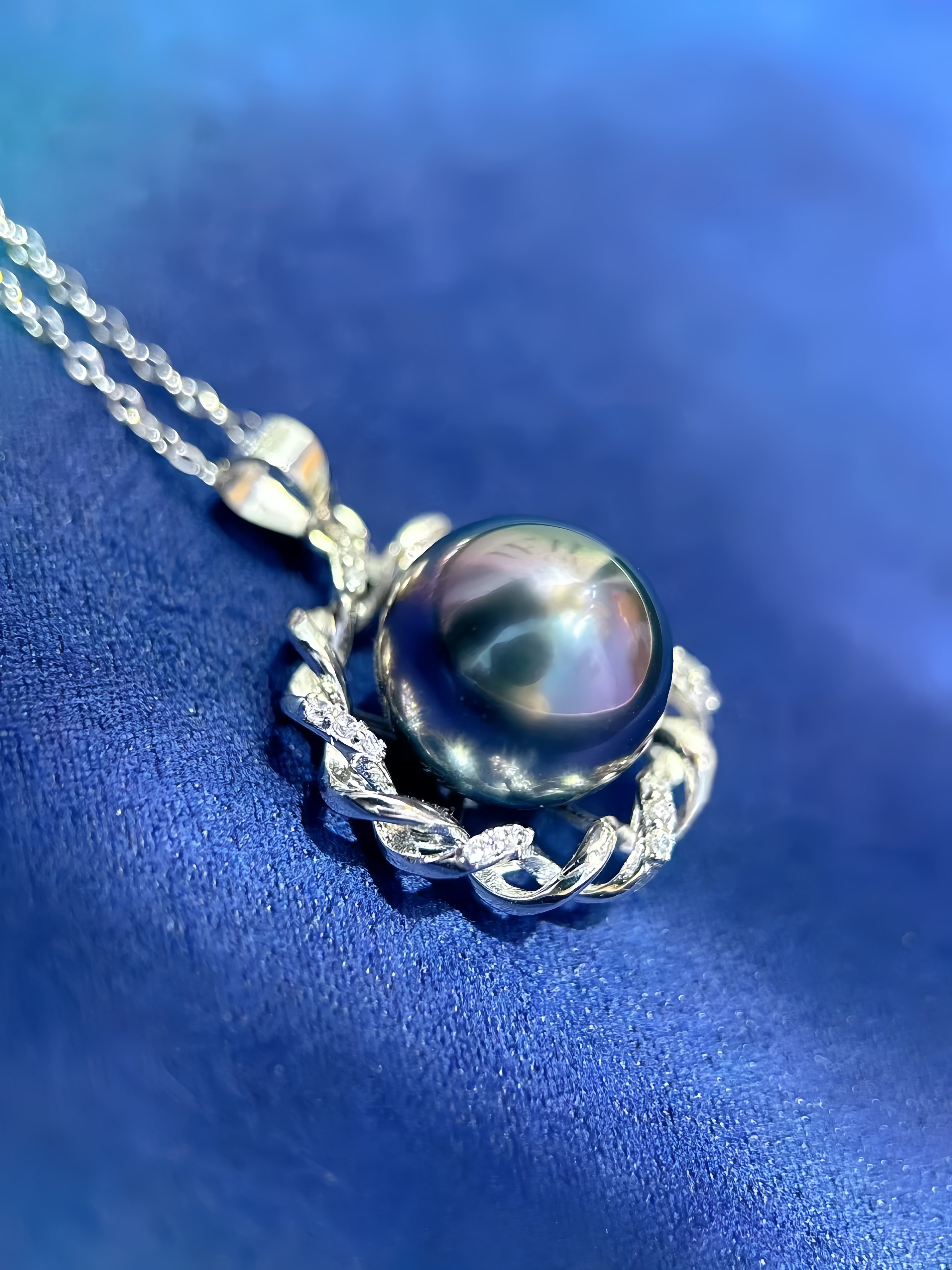 A close-up shot of a silver pendant necklace featuring a large, iridescent dark pearl surrounded by a textured silver wreath, resting on a blue fabric background.