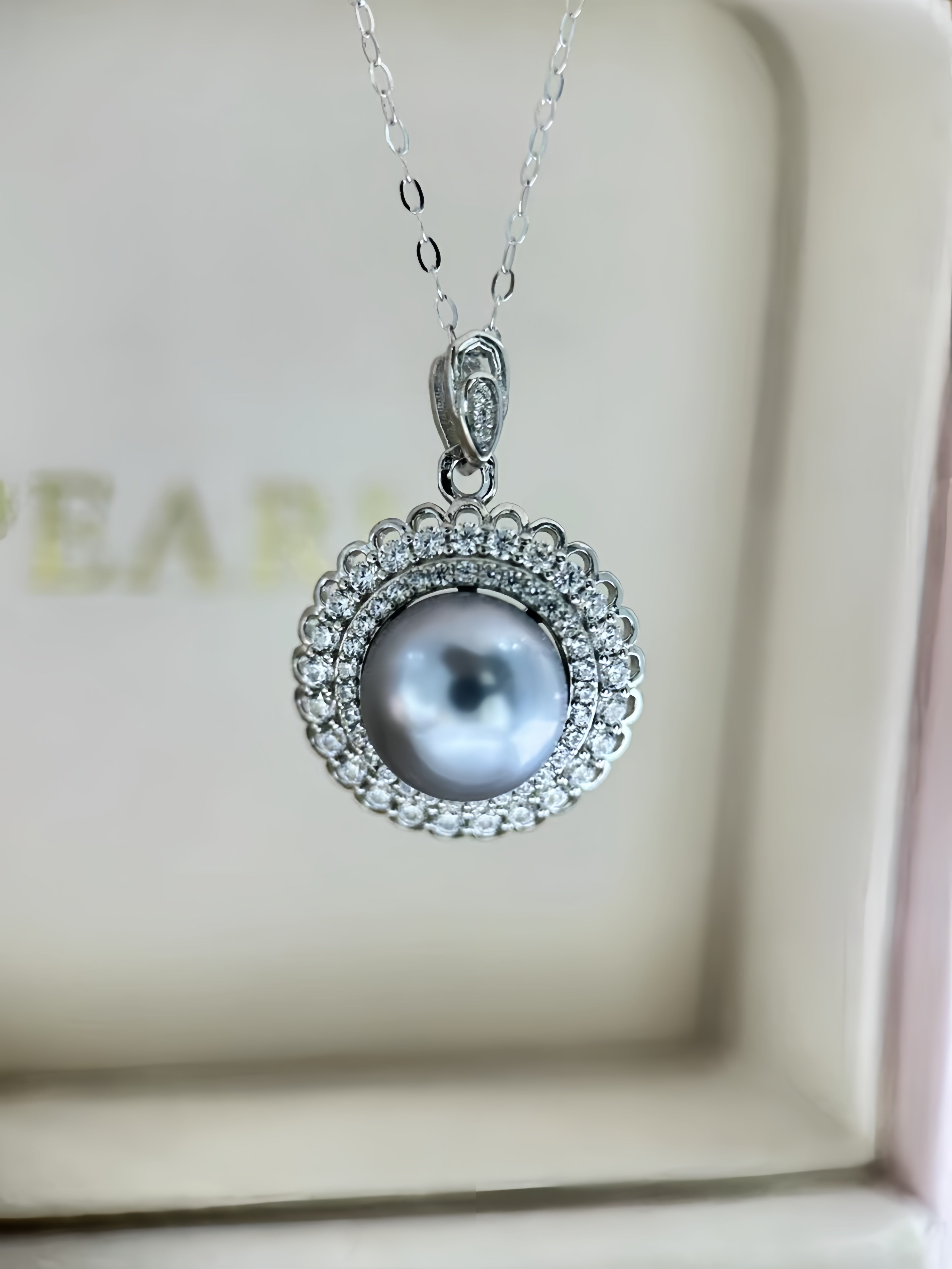 A silver pendant necklace featuring a central gray pearl surrounded by a halo of small clear stones, displayed inside a cream-colored jewelry box.