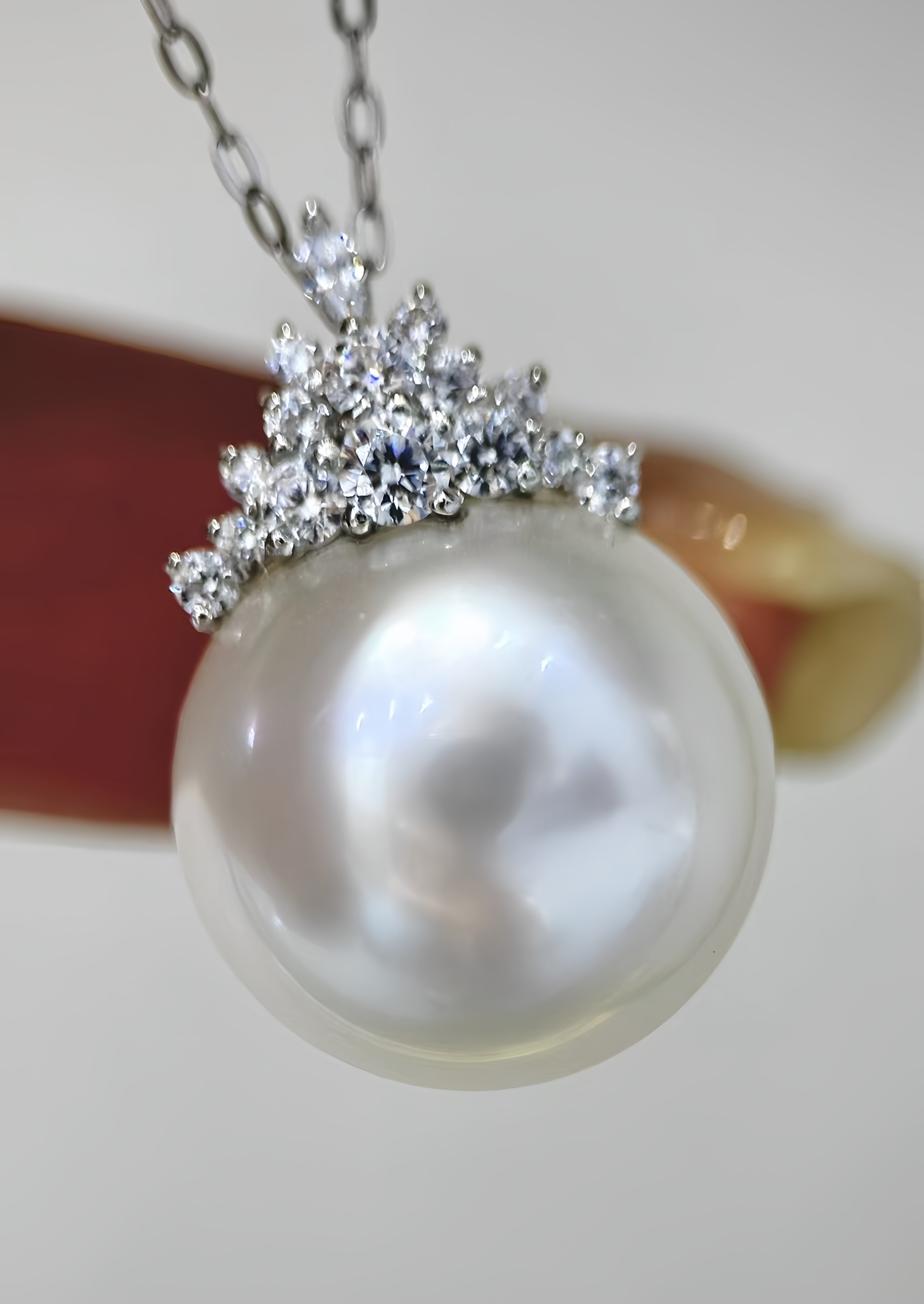 A close-up of a pearl pendant necklace with a silver-toned crown-like setting of clear stones, held against a soft neutral background.