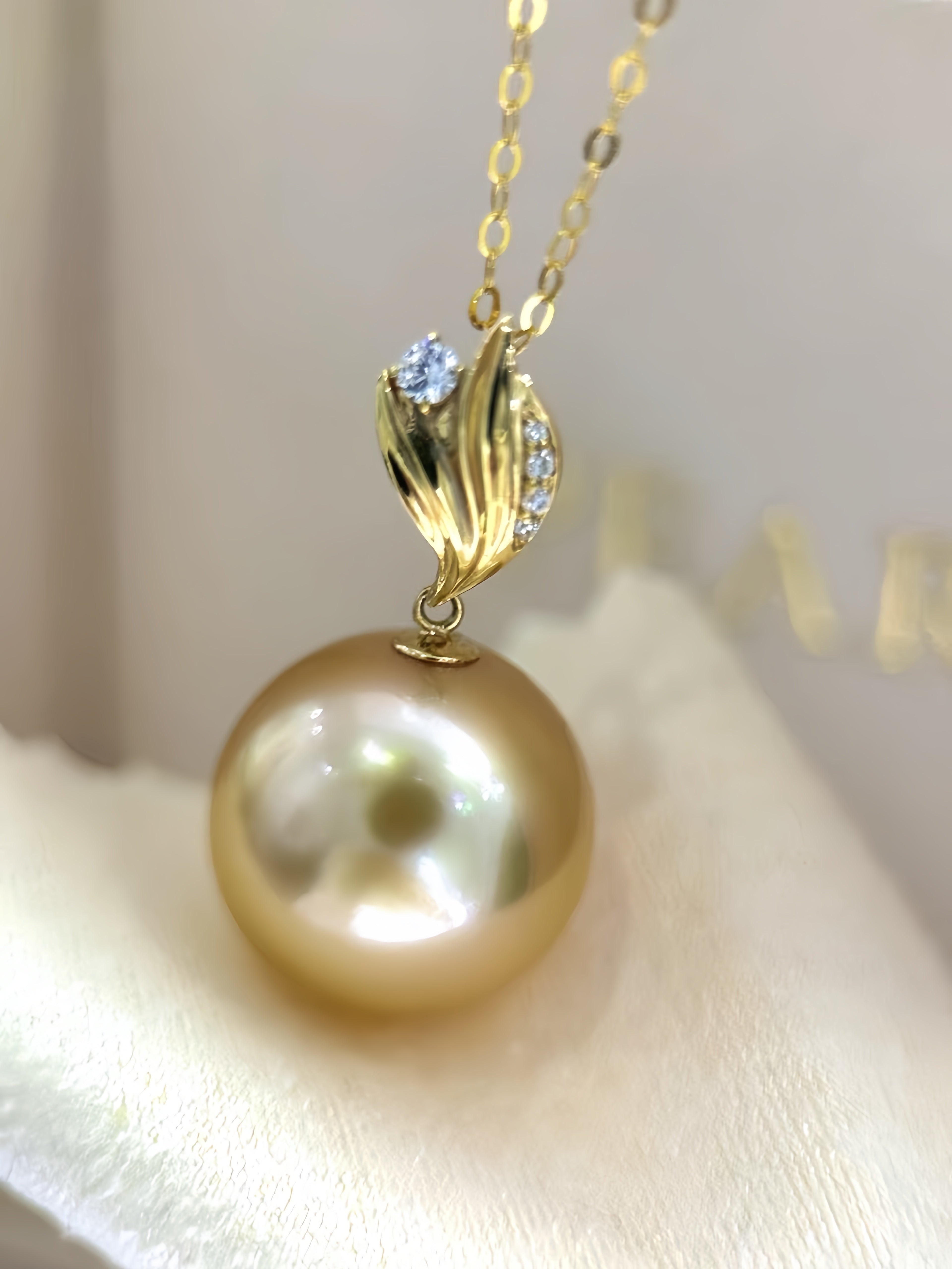 A gold pendant necklace featuring a large round freshwater pearl and a gold leaf-shaped bail accented with small clear stones, resting on a soft cream-colored fabric surface.