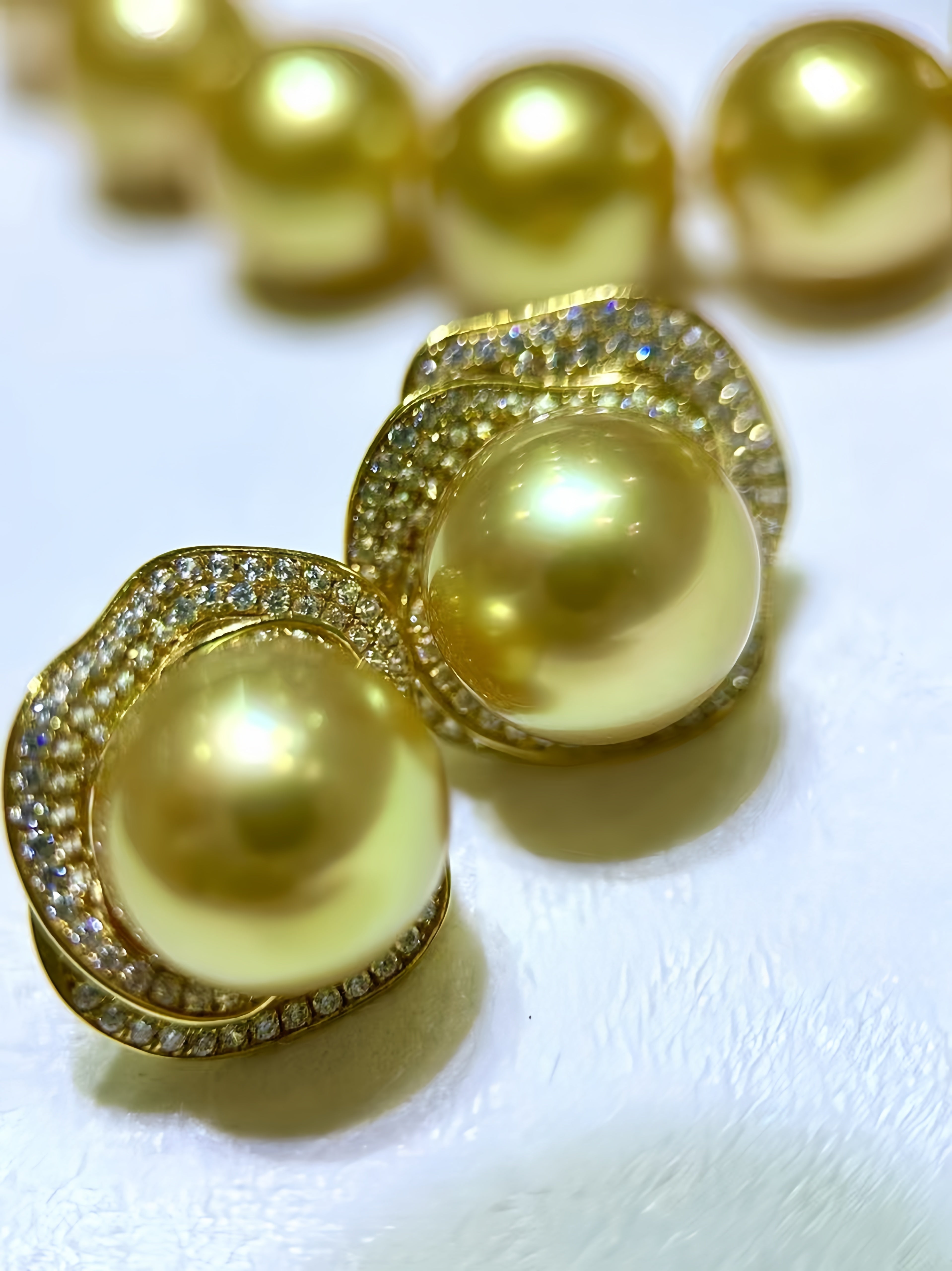 Gold-plated pearl stud earrings with a halo of small clear crystals, shown on a white textured surface with a blurred matching strand in the background.