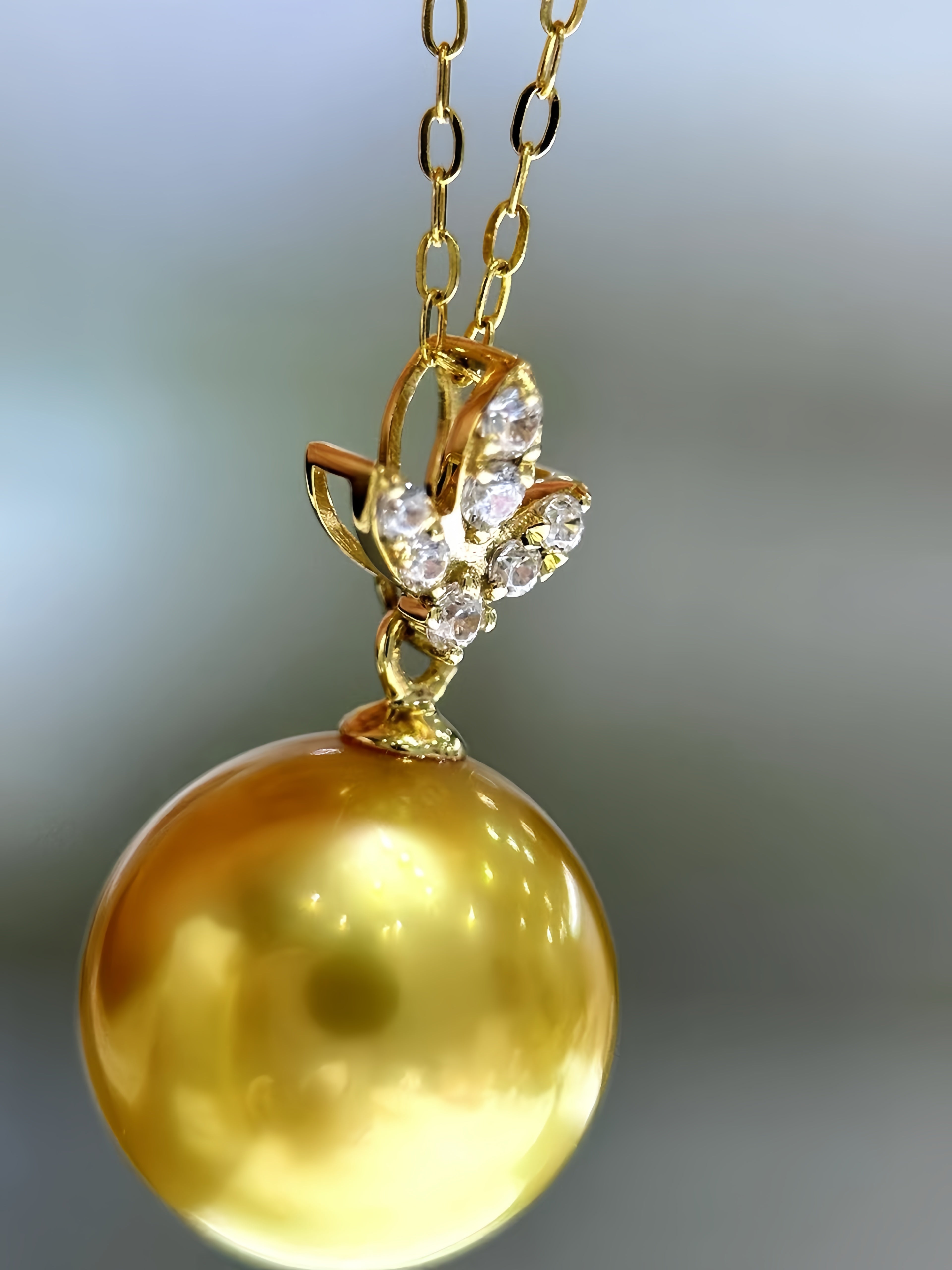 A close-up of a gold pendant necklace featuring a large, glossy round yellow-gold pearl suspended from a gold chain with a small crystal-accented setting.