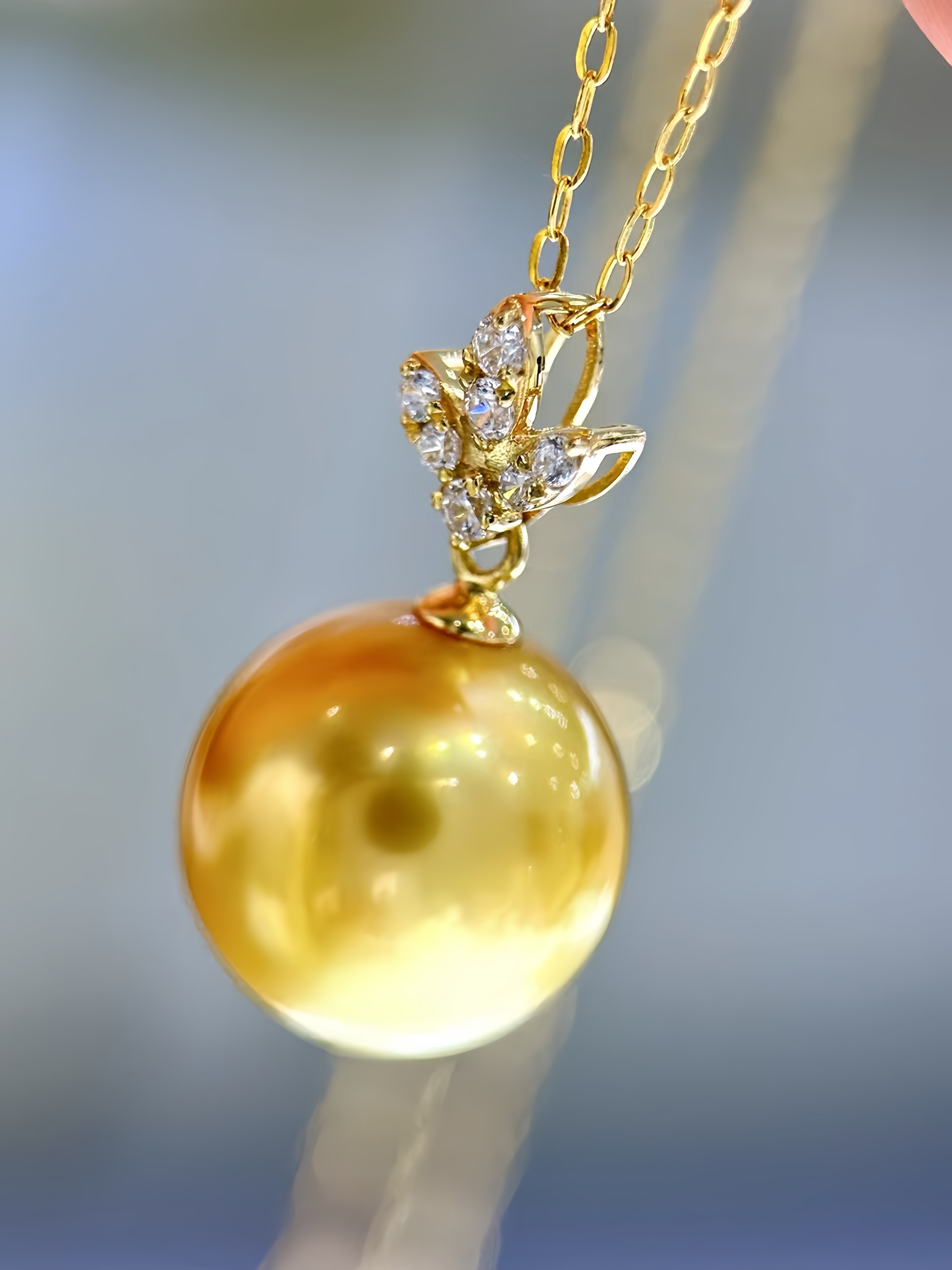 A close-up of a gold pendant necklace featuring a large, round, lustrous golden pearl suspended from a delicate gold chain. The pendant is adorned with a small gold leaf or branch design set with clear crystals, all set against a soft, blurred blue-gray background.