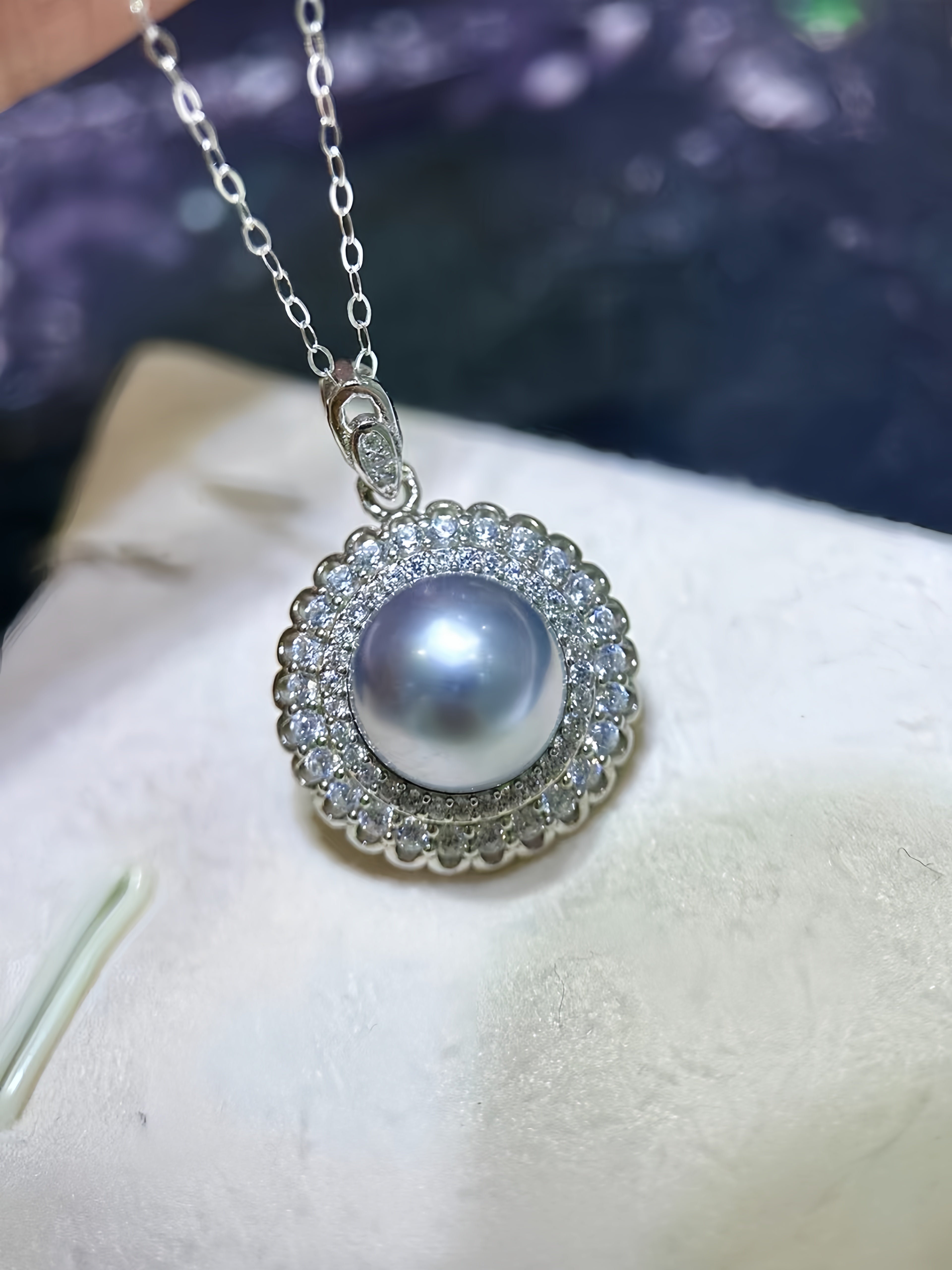 A silver pendant necklace featuring a large, lustrous gray pearl surrounded by a halo of small clear stones, resting on a textured white surface.