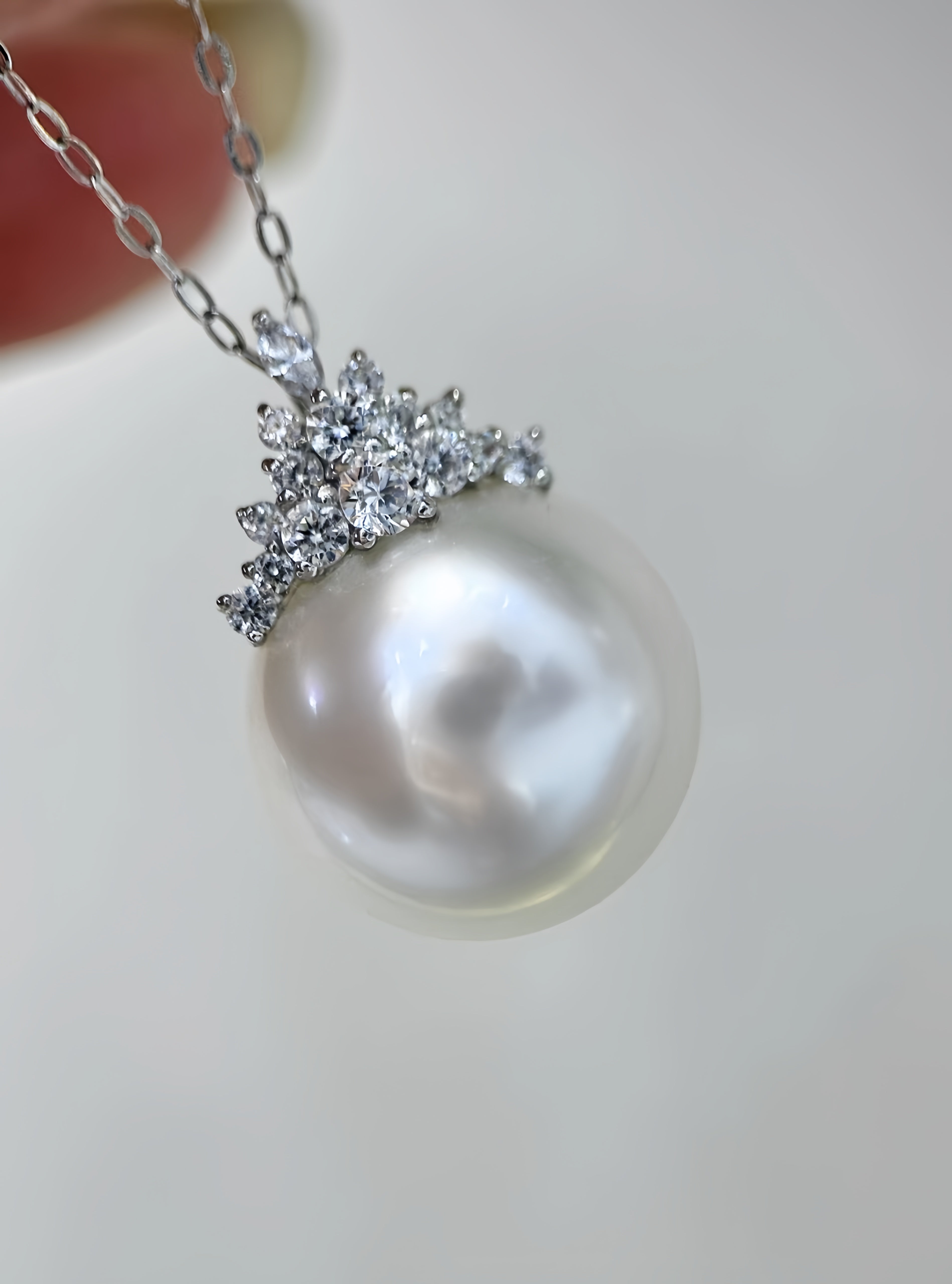 A close-up of a pearl pendant necklace with a silver-toned chain and a silver-toned crown-like setting adorned with small clear stones, held against a plain light background.