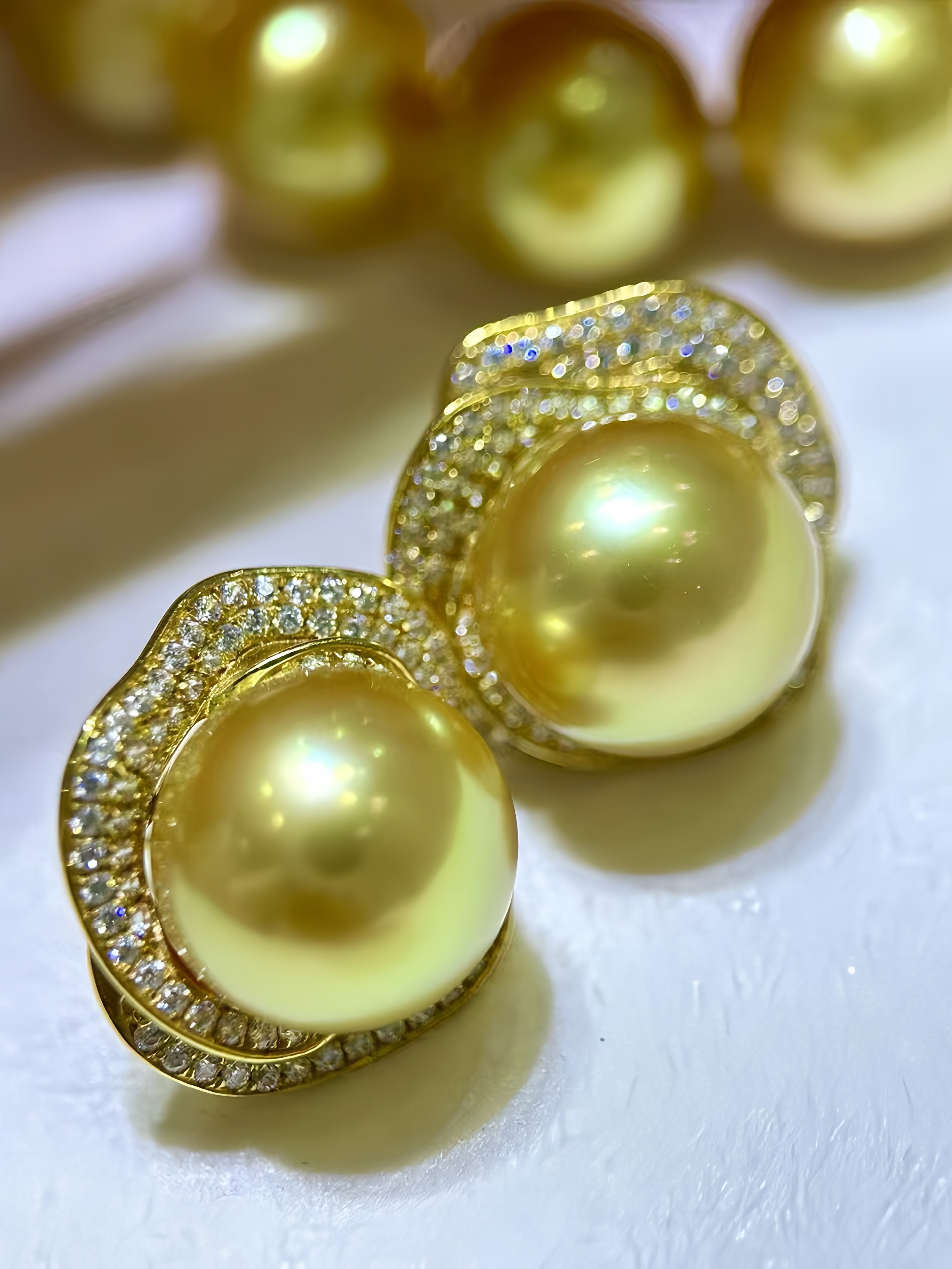 A pair of gold-toned stud earrings featuring large round yellow pearl-like centers surrounded by pavé-set clear stones, photographed on a light surface with additional blurred pearls in the background.