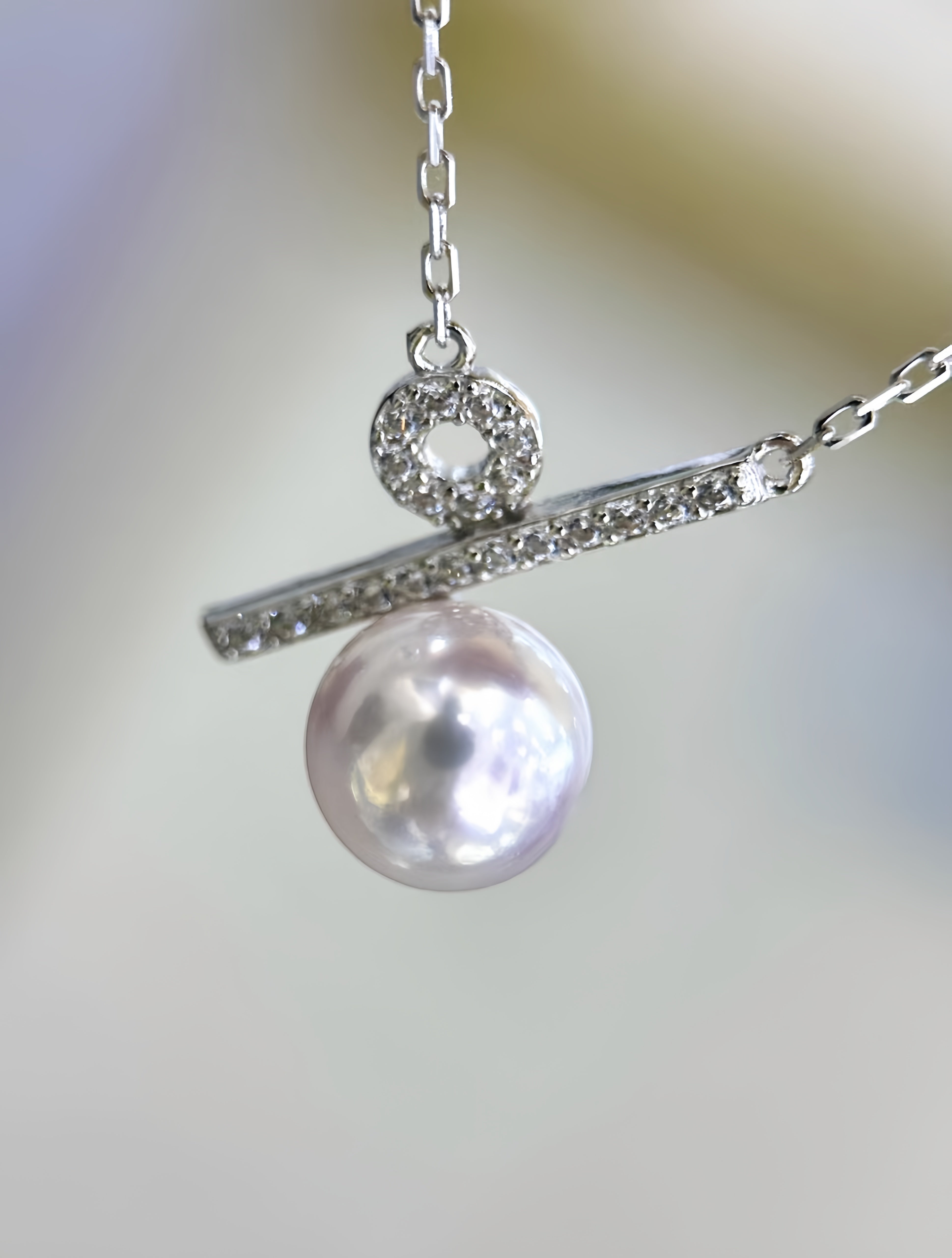 A close-up of a silver-toned necklace with a horizontal bar set with small clear stones, a circular ring detail, and a dangling lustrous pearl pendant, shown against a soft blurred light background.