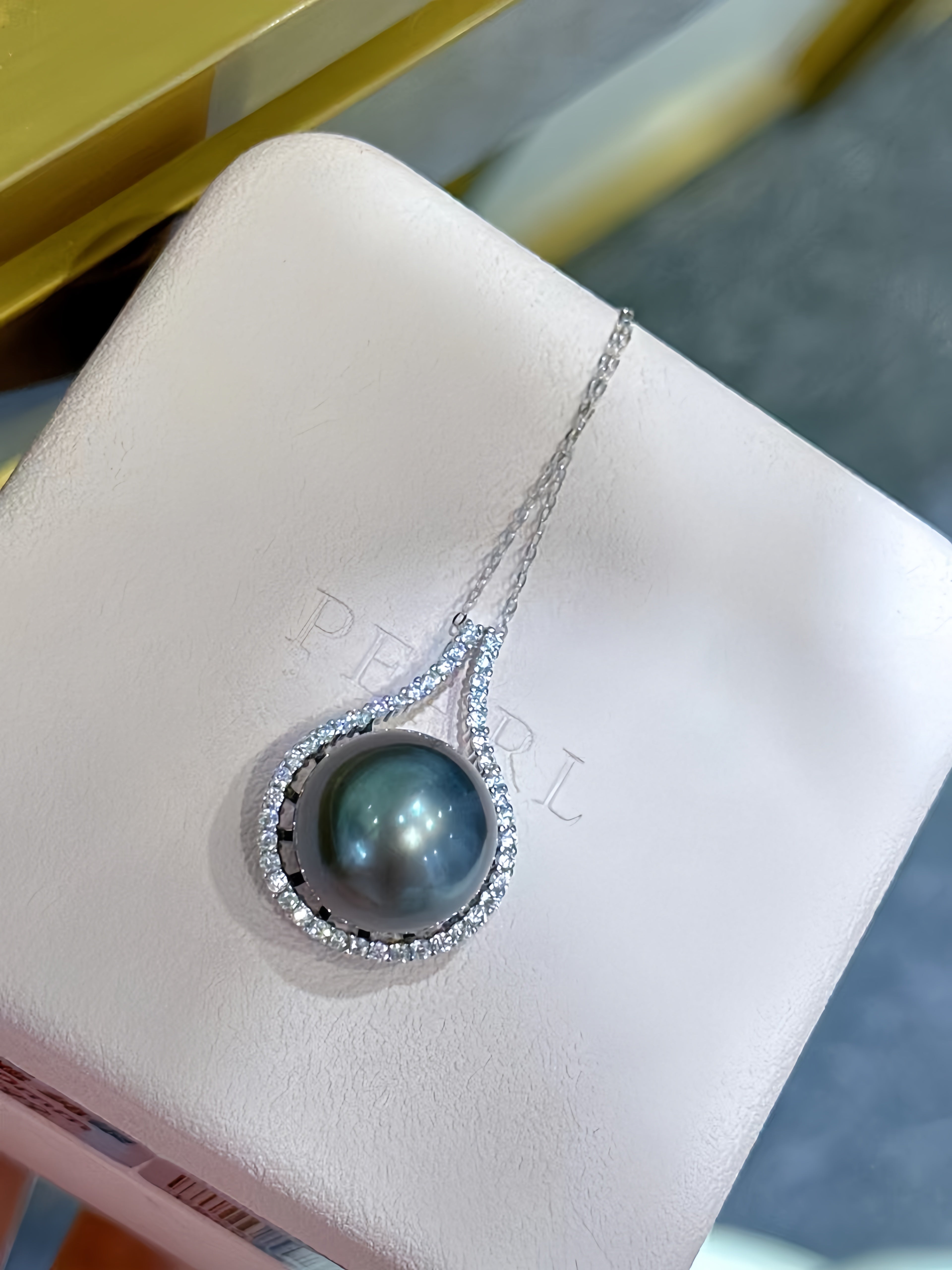 A teardrop-shaped pendant necklace featuring a dark gray iridescent freshwater pearl surrounded by a halo of small clear stones, displayed on a white jewelry box.