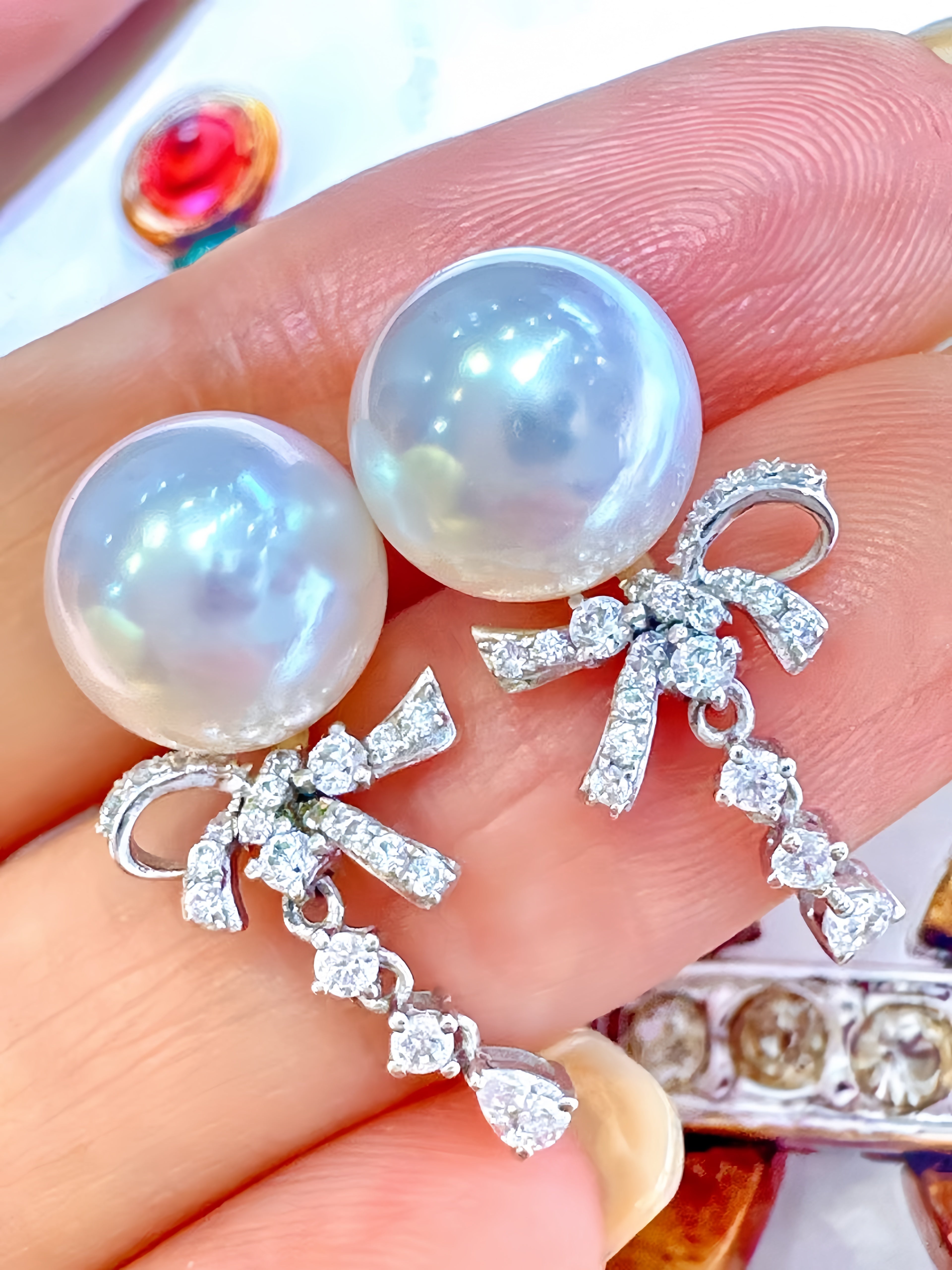 A pair of pearl and crystal bow drop earrings held on a person's fingers. The earrings feature large, lustrous white pearls at the top, connected by a silver-toned bow embellished with clear crystals, with dangling crystal accents below.
