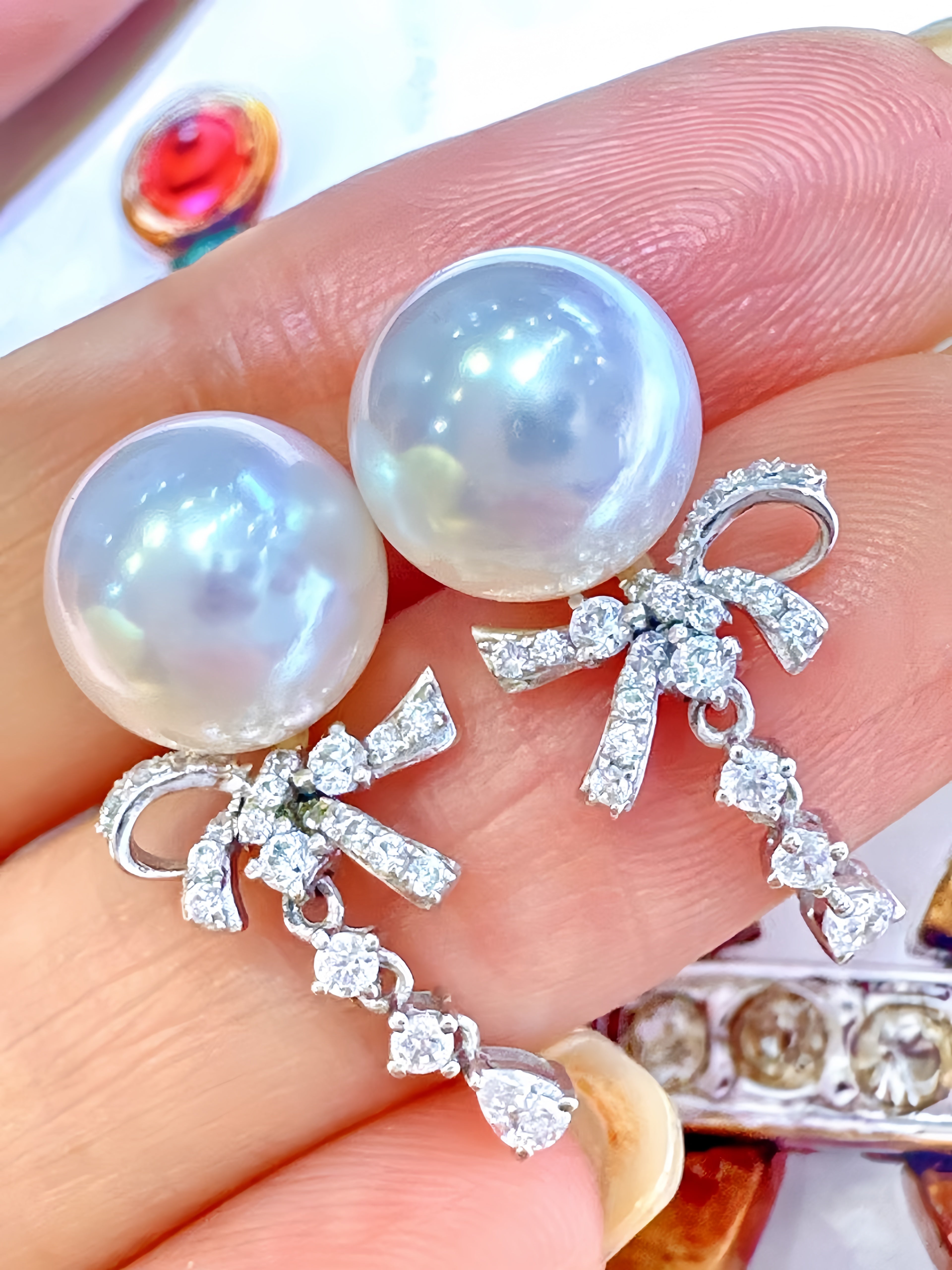 A pair of pearl and silver bow drop earrings with clear crystal accents, held on a person's fingers.