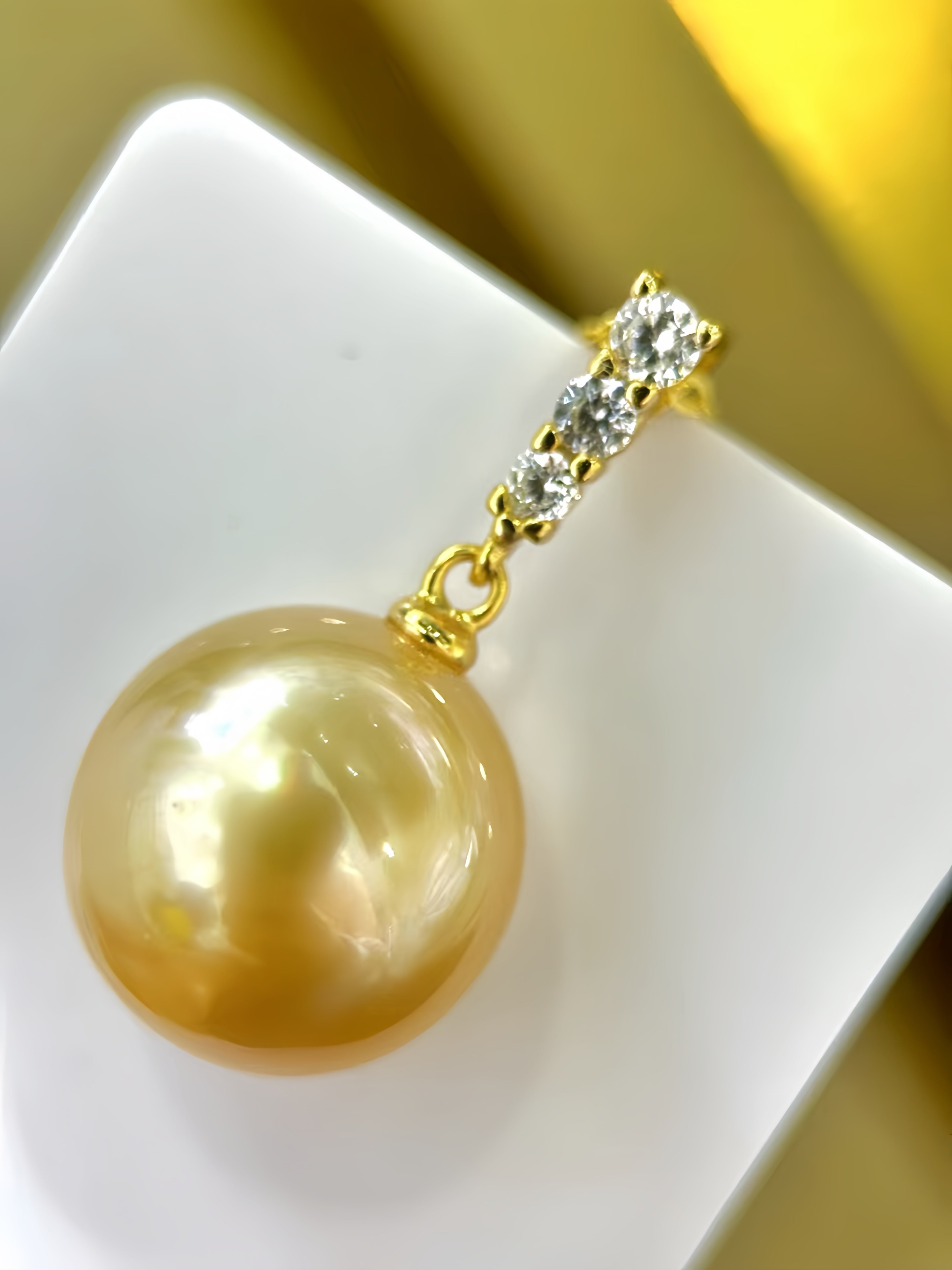 A gold pendant featuring a large, lustrous golden pearl and a small cluster of clear stones, displayed on a white geometric stand.
