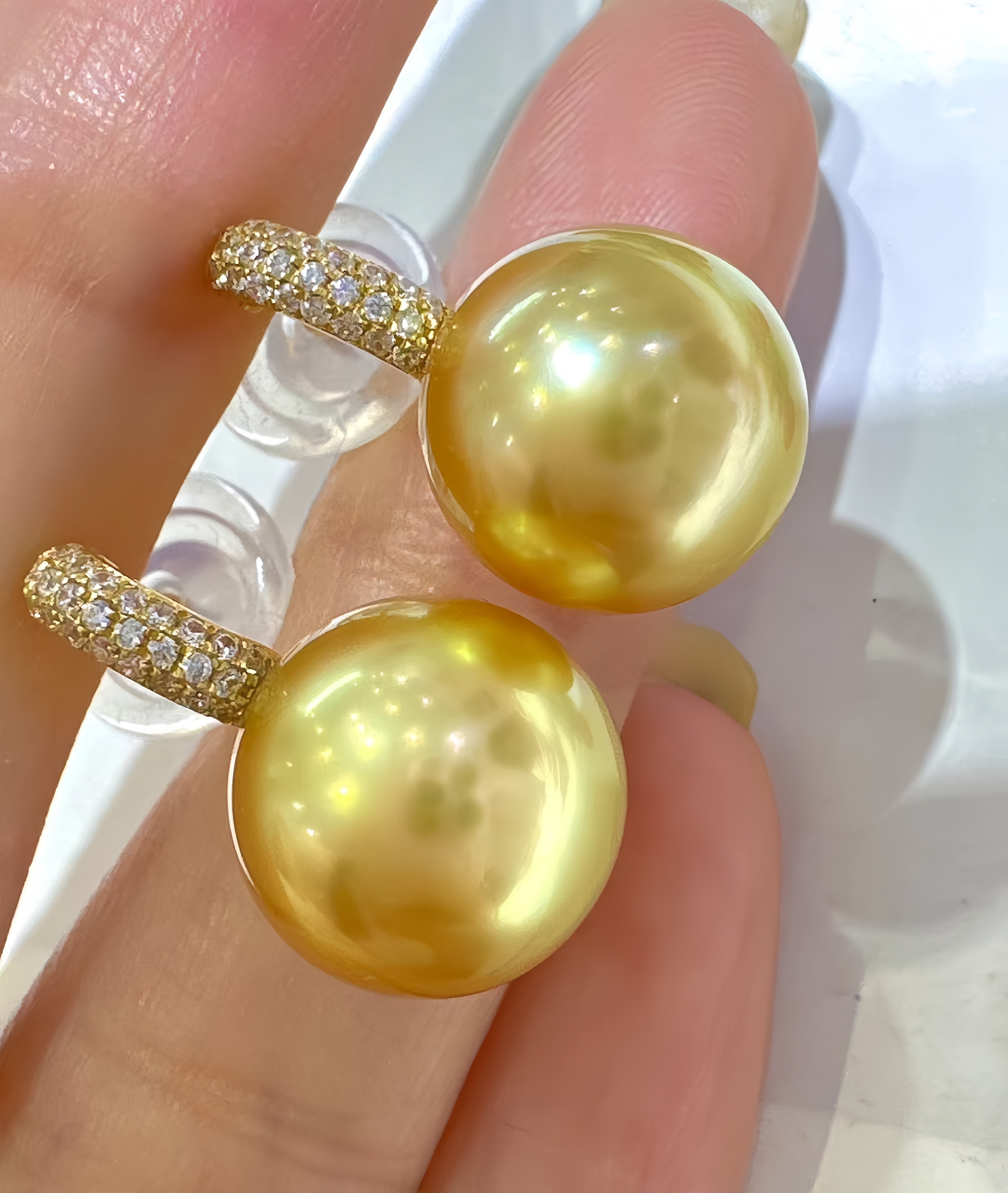 A pair of gold hoop earrings with large round yellow pearl drops, held between fingers. The hoops are paved with small clear stones.