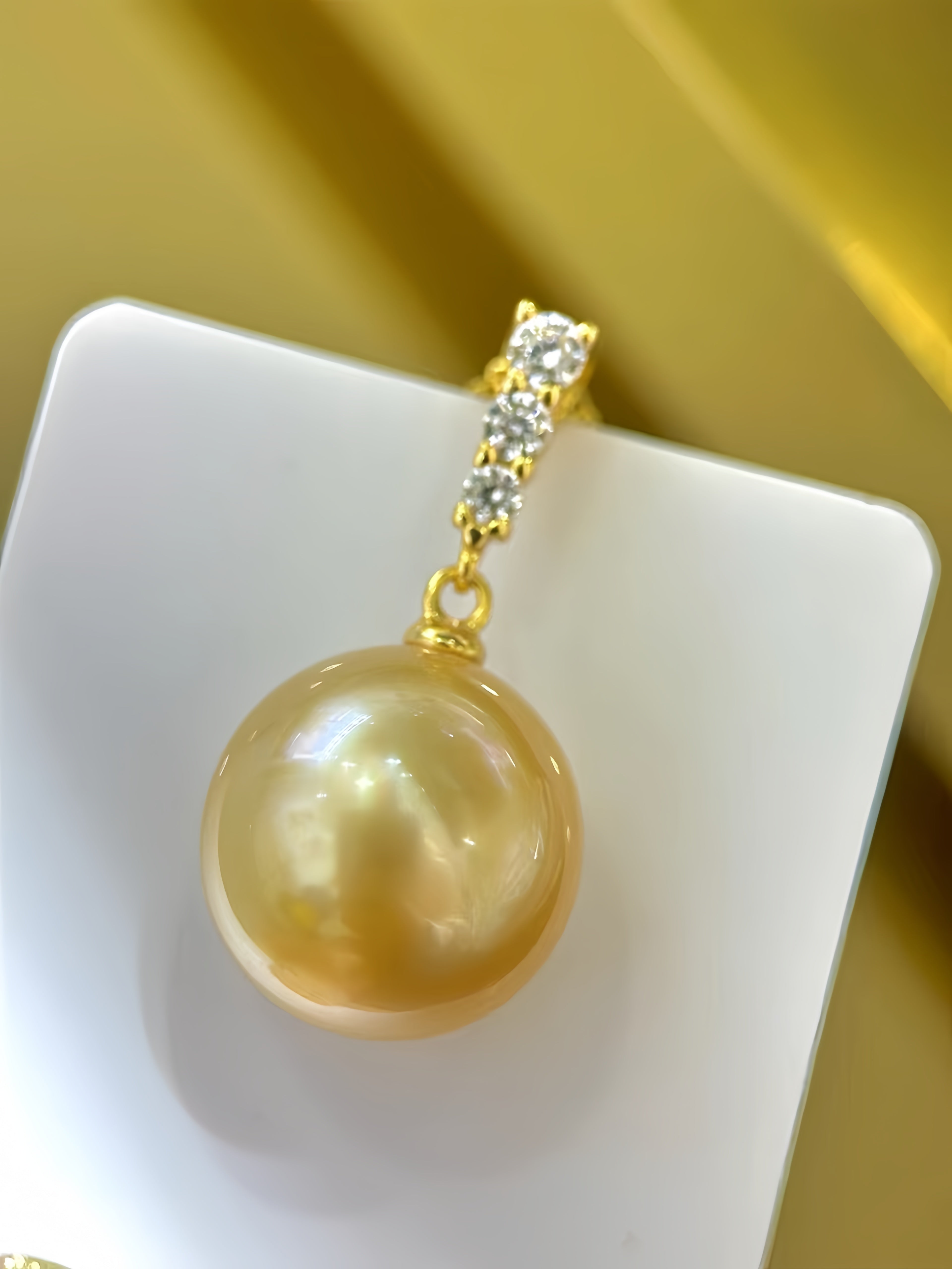 A gold pendant featuring a large, lustrous golden pearl and a small cluster of clear stones at the top, displayed on a white square card against a gold background.