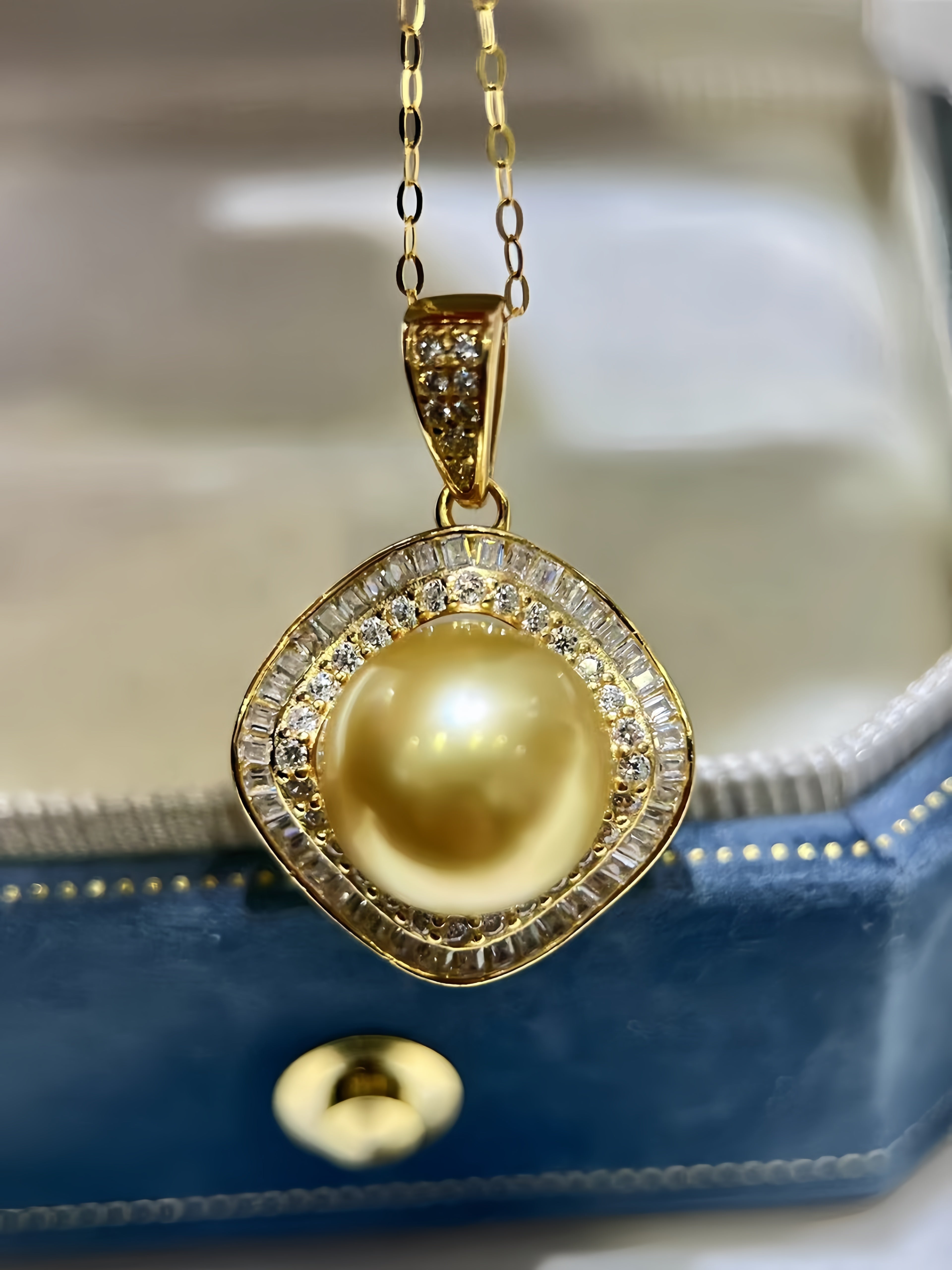 A gold pendant necklace featuring a large round yellow pearl surrounded by a halo of small clear stones, resting on a blue jewelry box.