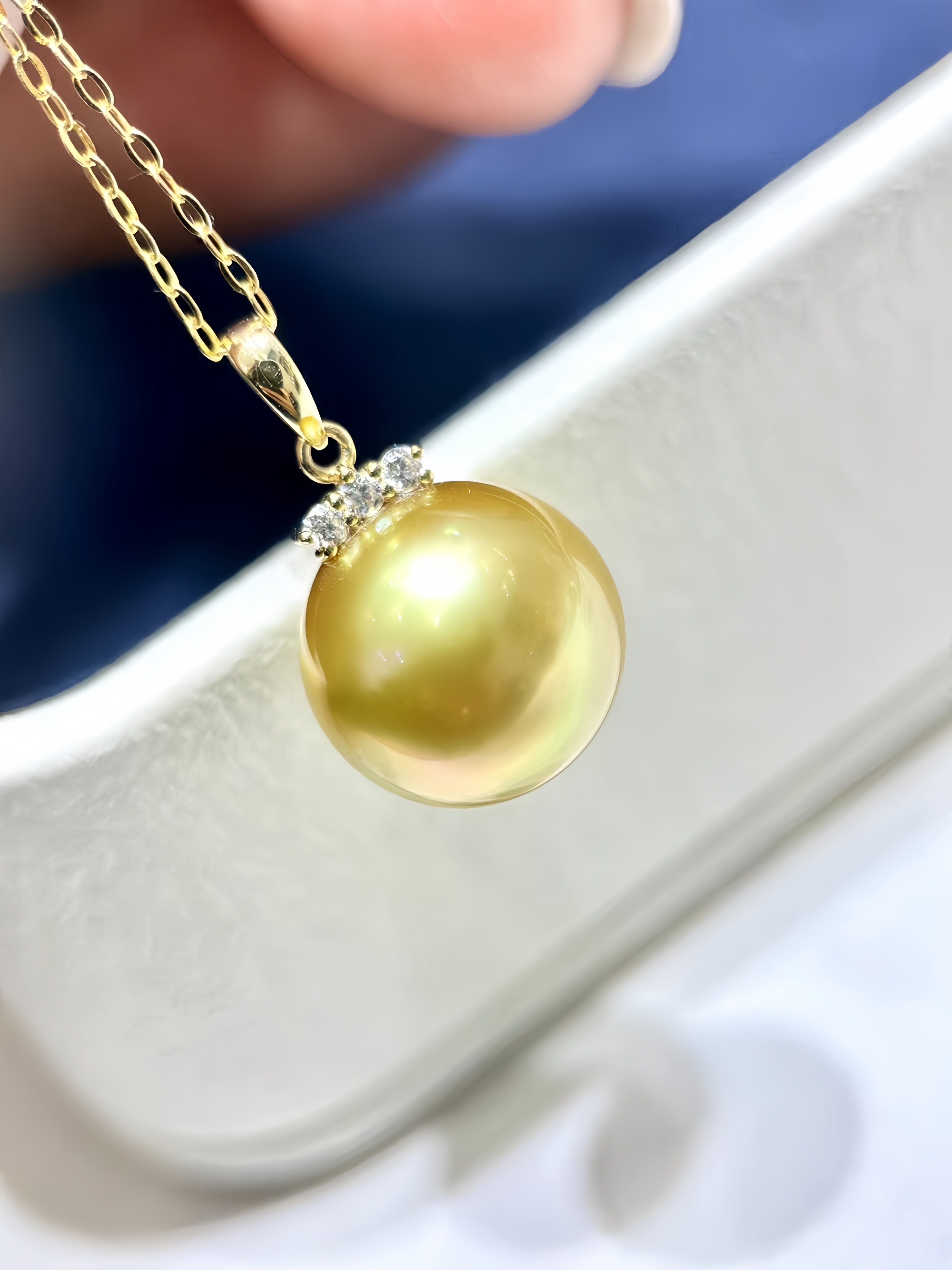 A close-up of a gold chain necklace with a round, lustrous yellow-gold pearl pendant, held near a white jewelry box. The pendant is set with small clear stones at the top.
