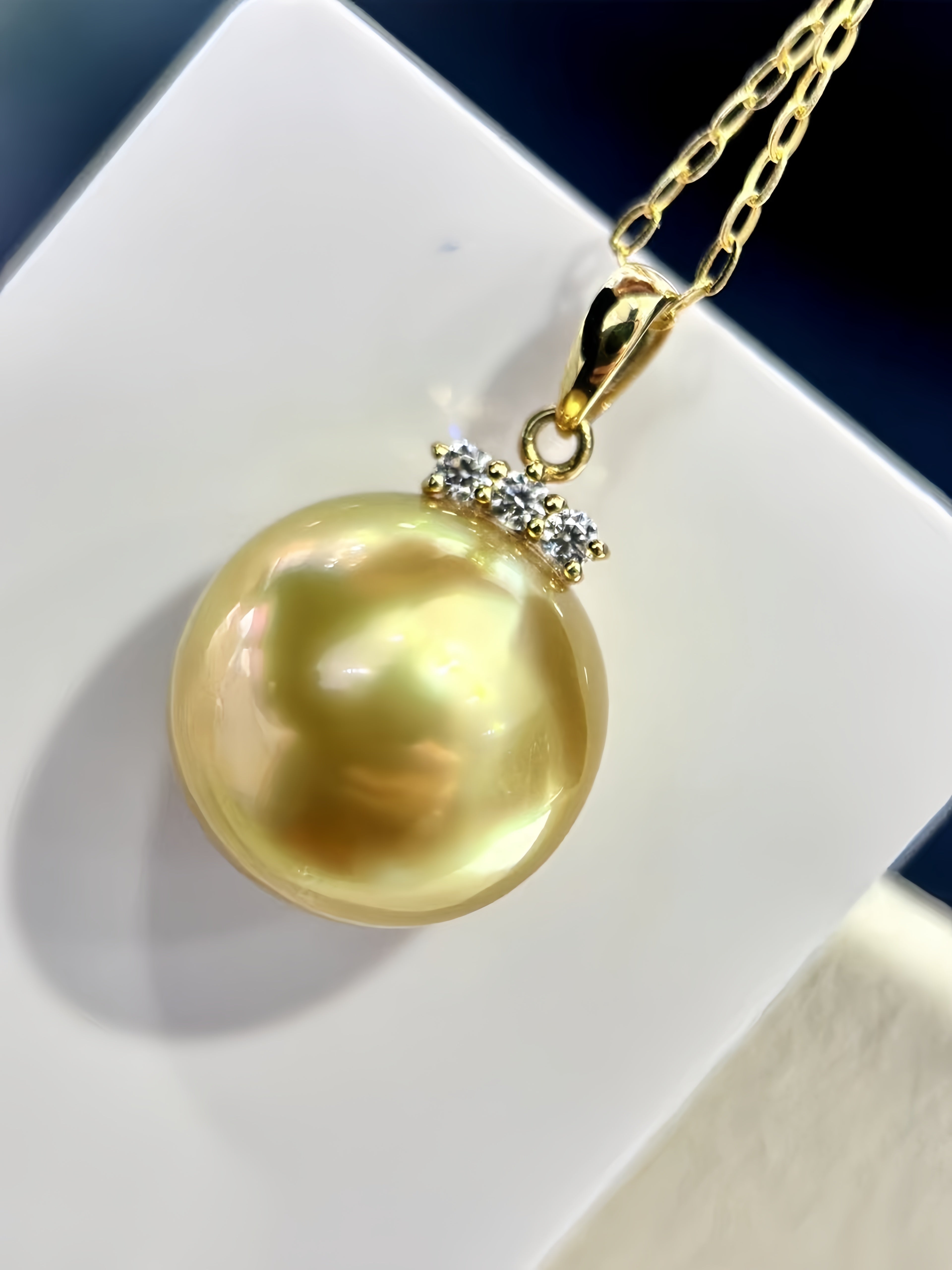 A gold pendant necklace featuring a large, lustrous yellow pearl with a small cluster of clear stones at the top, displayed on a white geometric block.