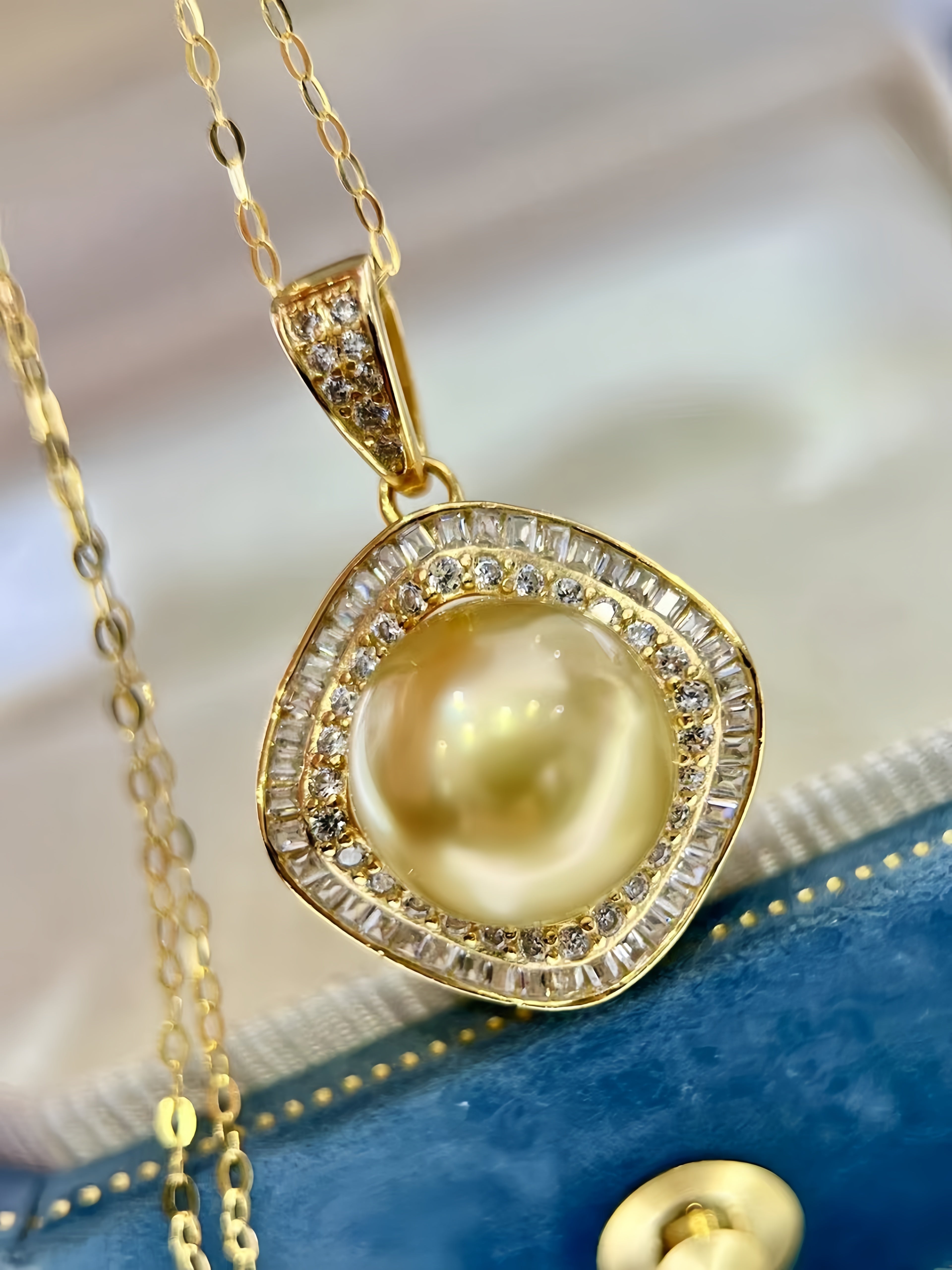 A gold pendant necklace featuring a large round cream-colored pearl surrounded by a double halo of small clear stones, displayed on a blue jewelry box.