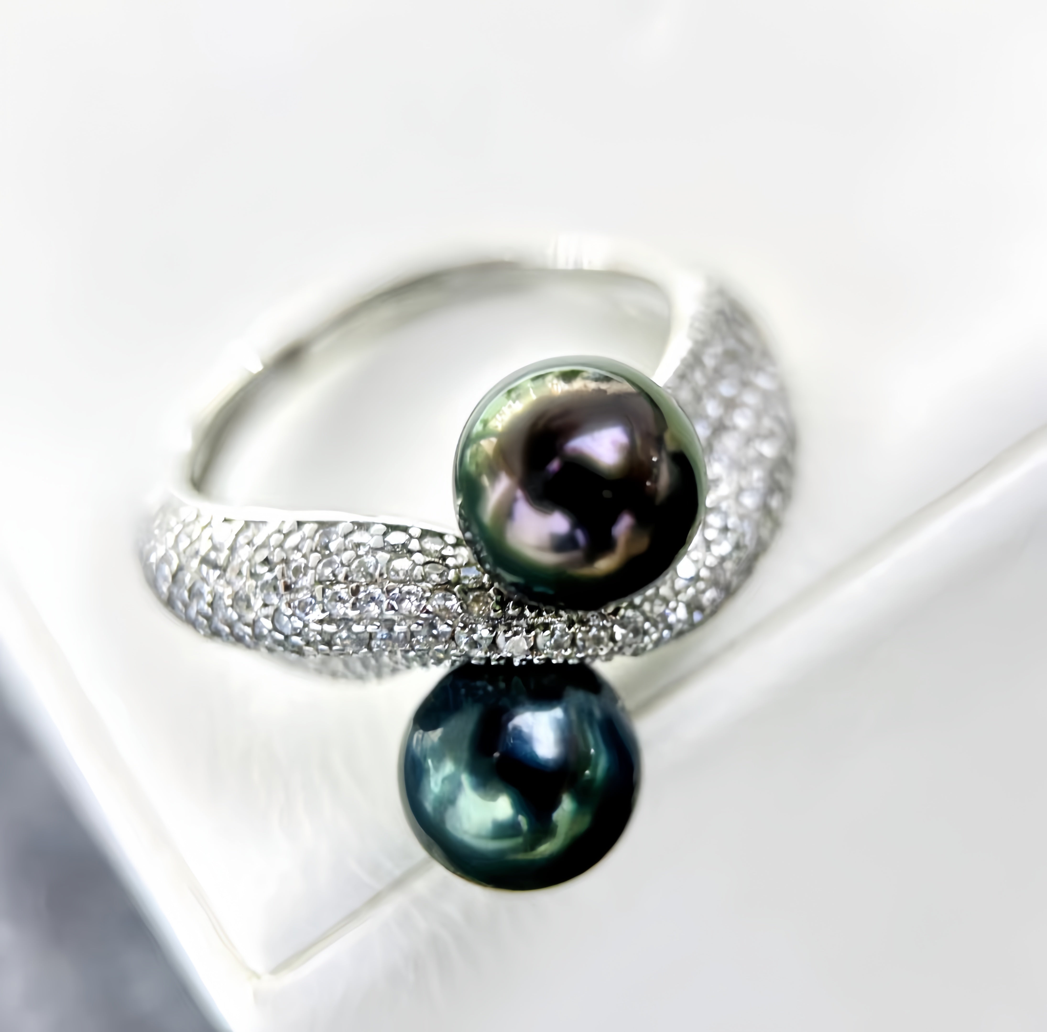 A silver ring featuring two dark green-black pearls and a pavé-set silver band, displayed on a white jewelry stand.
