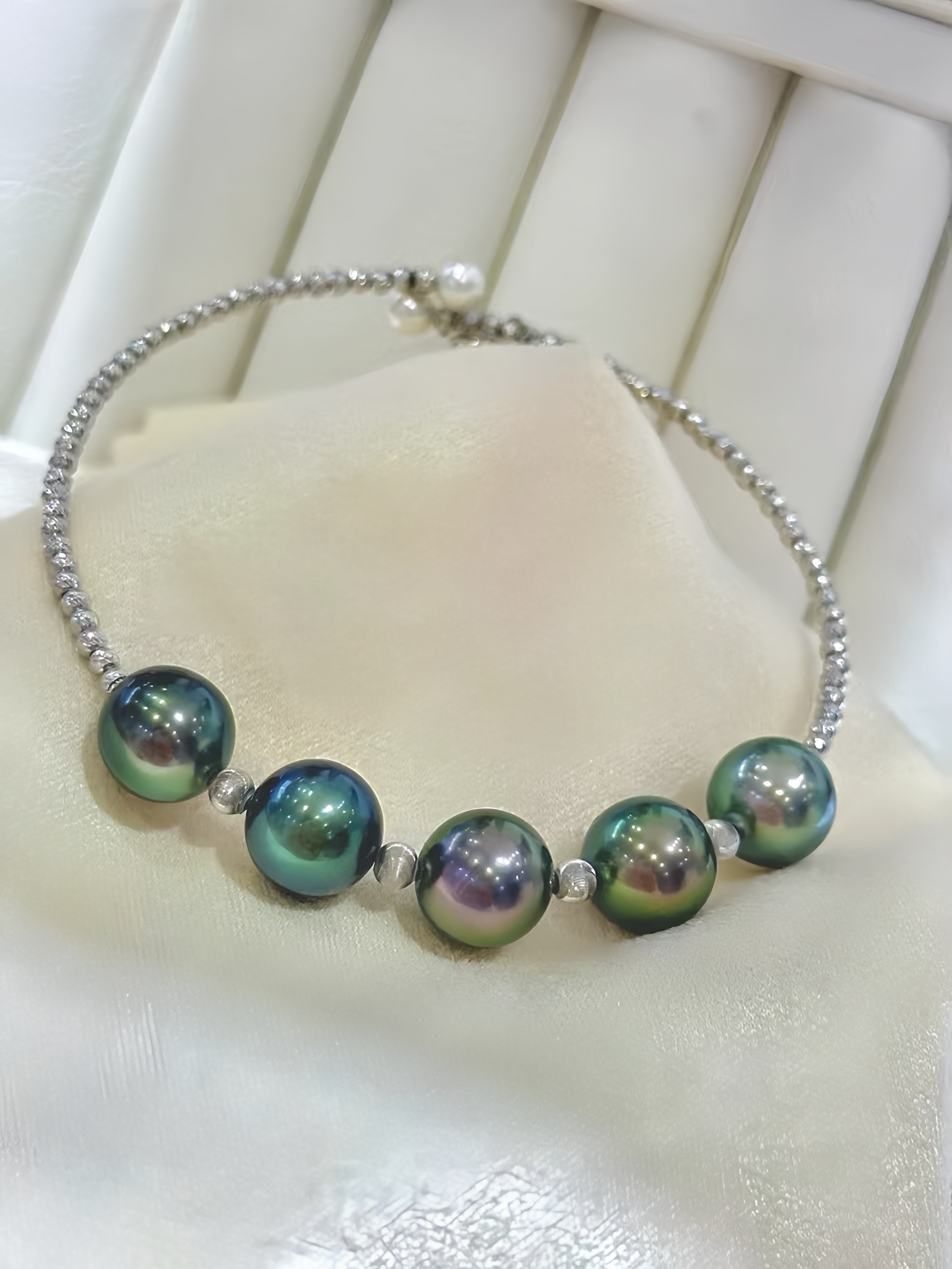 A silver beaded chain bracelet featuring five large, iridescent green and blue pearls separated by small silver spacer beads, displayed on a cream-colored jewelry stand.
