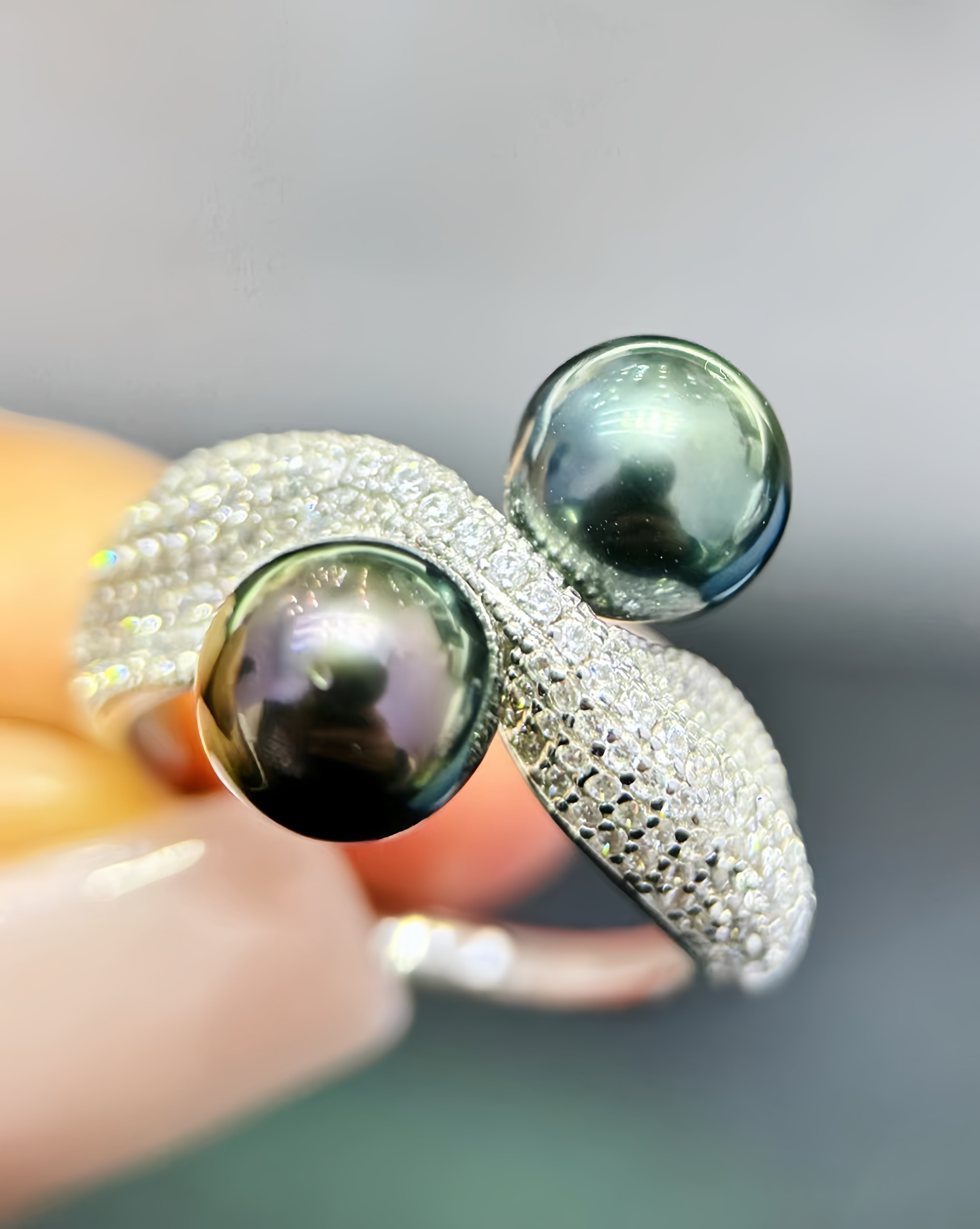 A close-up of a silver ring with two large dark green-black pearls, held by a hand. The band is heavily set with small clear crystals.