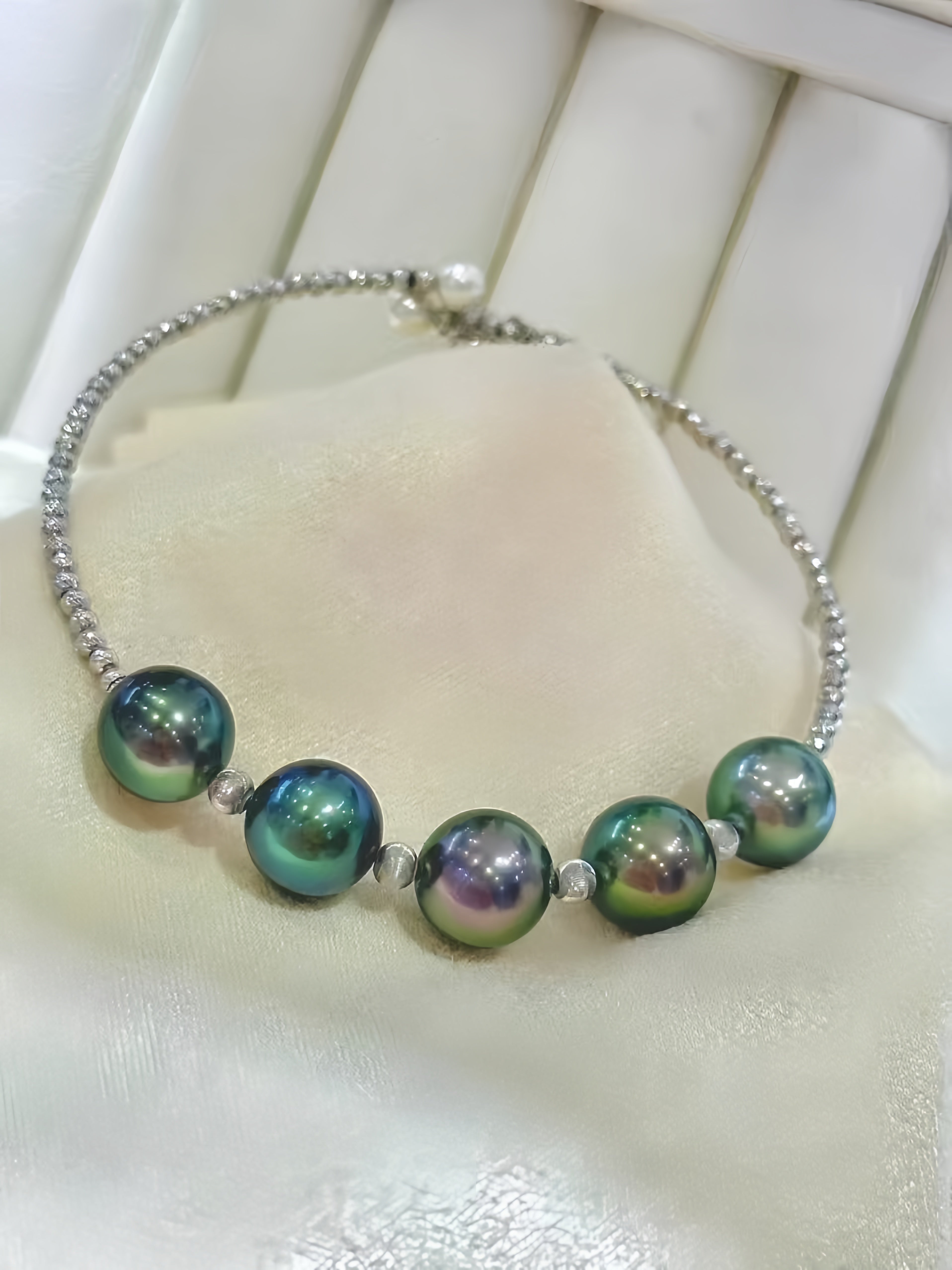 A silver beaded chain bracelet featuring five large, iridescent green and blue pearls, displayed on a cream-colored jewelry stand.