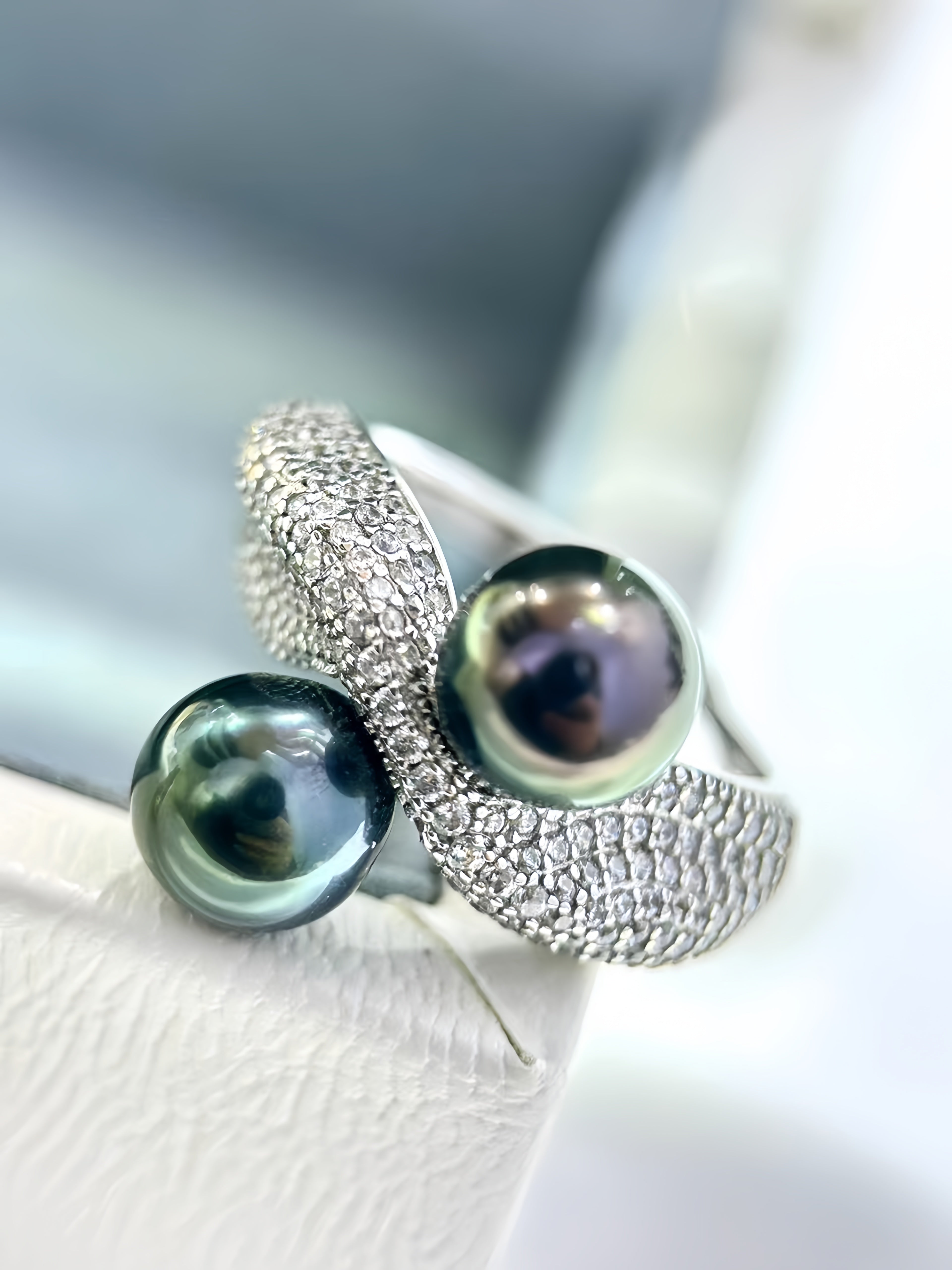 A silver ring featuring two large iridescent dark green pearls and a textured pavé band, resting on a white textured surface.